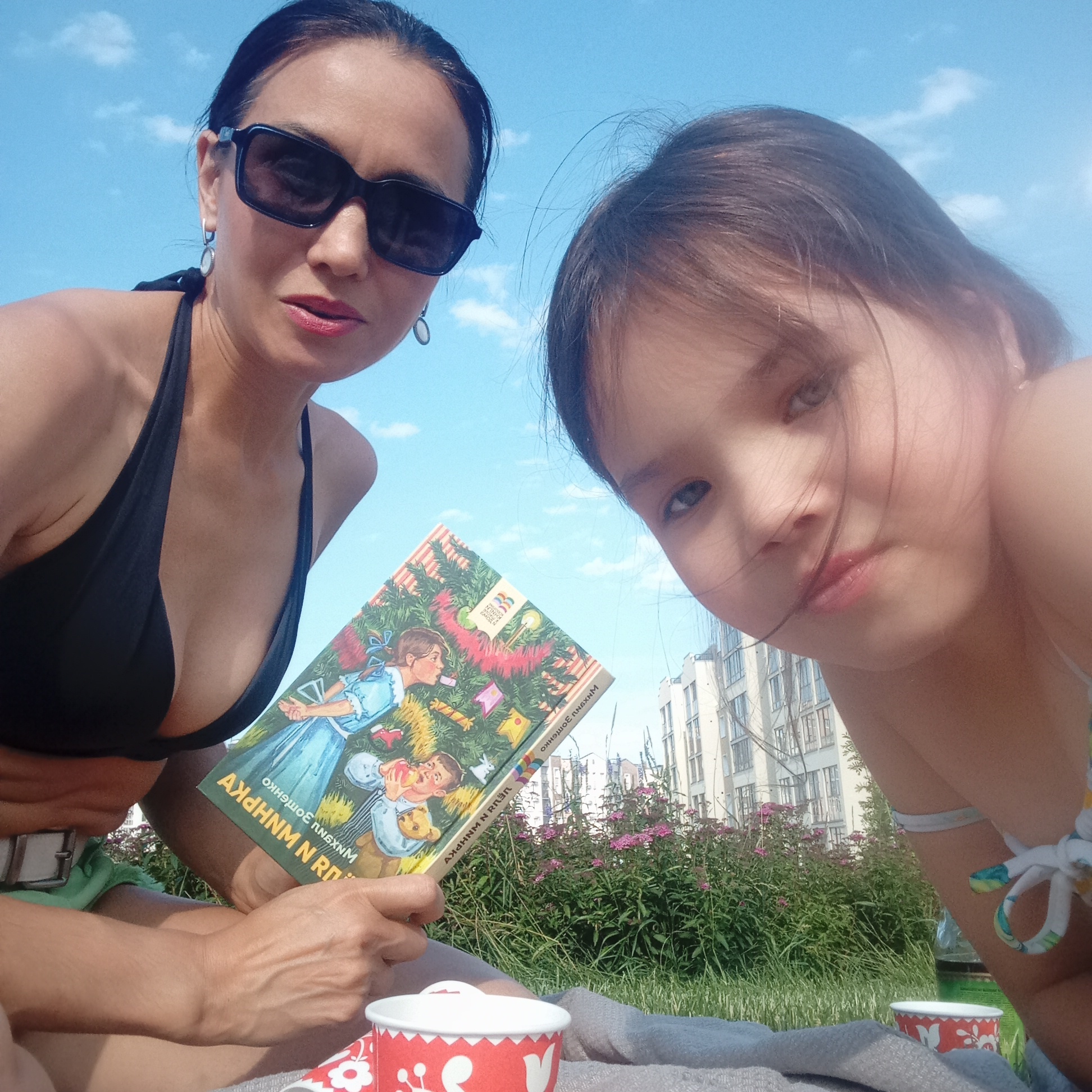 In a close-up selfie, two females are outdoors, with a bright blue sky and scattered clouds above. The female on the left, wearing sunglasses and a black bikini top, holds a book with an illustration of children decorating a Christmas tree. Her left arm is visible, resting on her leg. To the right, a younger female is partially visible, her head and shoulder in the foreground. In the background, lush green bushes with small pink flowers are in bloom, with a modern, multi-story building behind them. A red and white patterned cup sits on a grey blanket in the foreground. The scene appears to be set in a park or grassy area within the city of Pervomaysky, Russia.