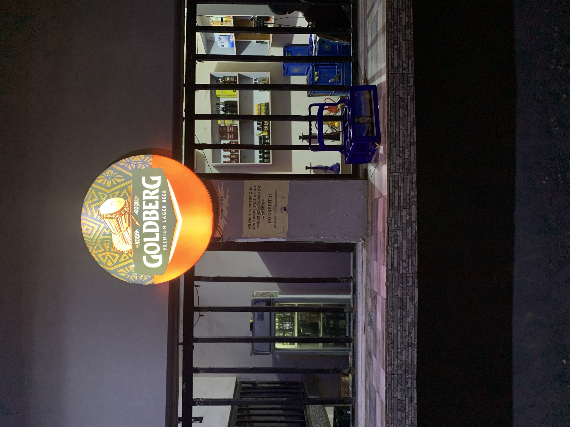 The image captures a section of a building in New Stadium, Nigeria, with a prominently displayed "GOLDBERG Premium Lager Beer" sign illuminated against a dark background. Behind the sign, a barred window reveals shelves stocked with bottles, possibly alcoholic beverages, suggesting a bar or store. Blue crates are visible, possibly for beer bottles, and a blue bottle carrier is placed near the window. A notice on the wall reads: "WE WISH TO NOTIFY OUR CUSTOMERS THAT WE NO LONGER SE DRINKS ON CREDIT...NO CREDIT!!! WE REGRET ANY ANVENIENCES". The environment appears to be outdoors at night, with a portion of the street visible on the right side of the frame. A refrigerator can be seen through the bars in the background.