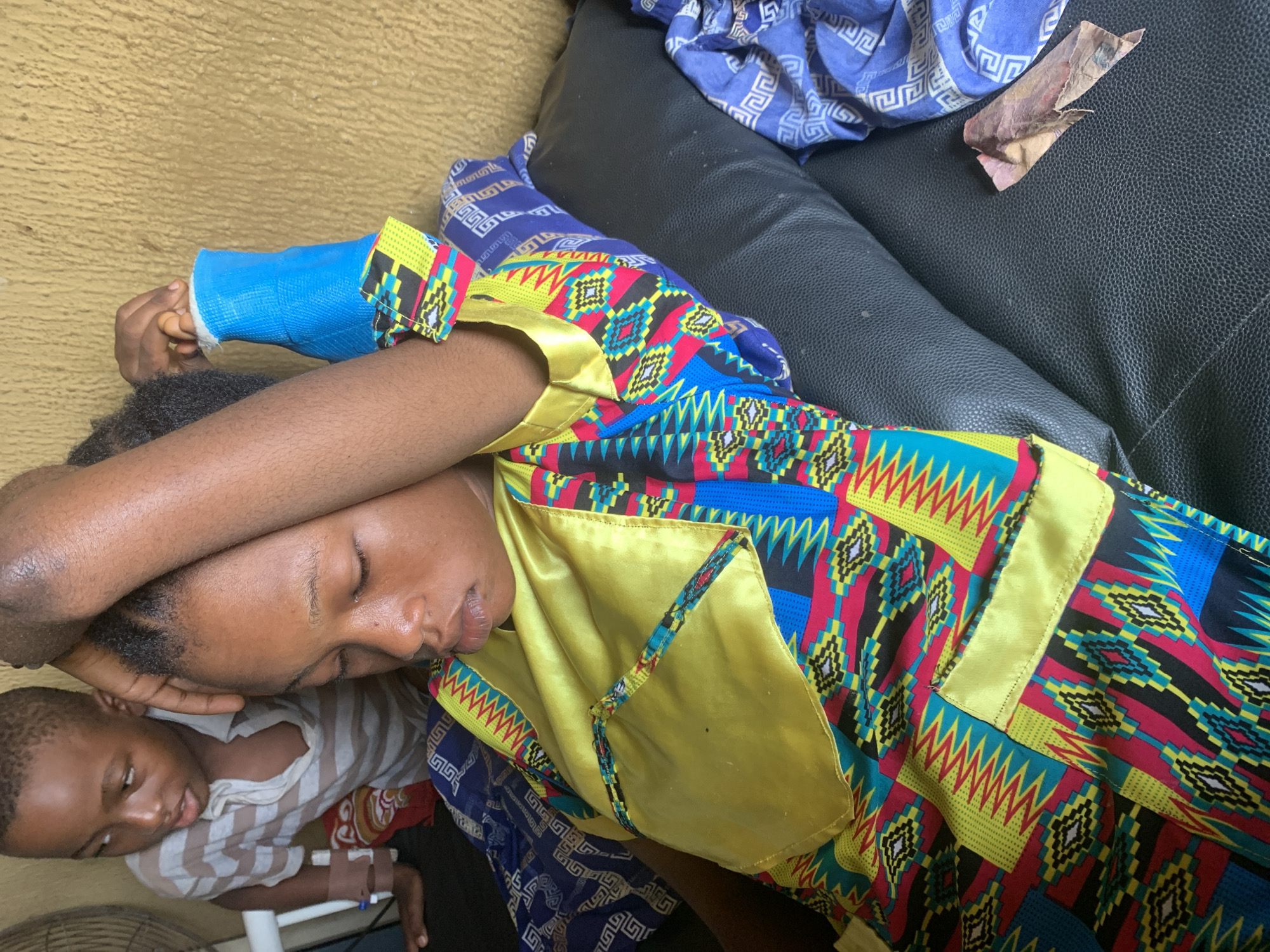 A young woman with closed eyes, wearing a brightly colored African print dress and a blue cast on her left arm, rests her head on her arm, which is raised above her head. She is lying down on a black leather surface. A young boy in a striped t-shirt stands behind her, looking towards the viewer and slightly to the left. He has a medical bracelet on his left wrist. The background consists of a textured, light brown wall. The scene appears to be indoors, likely in a home environment in Shagamu, Nigeria.