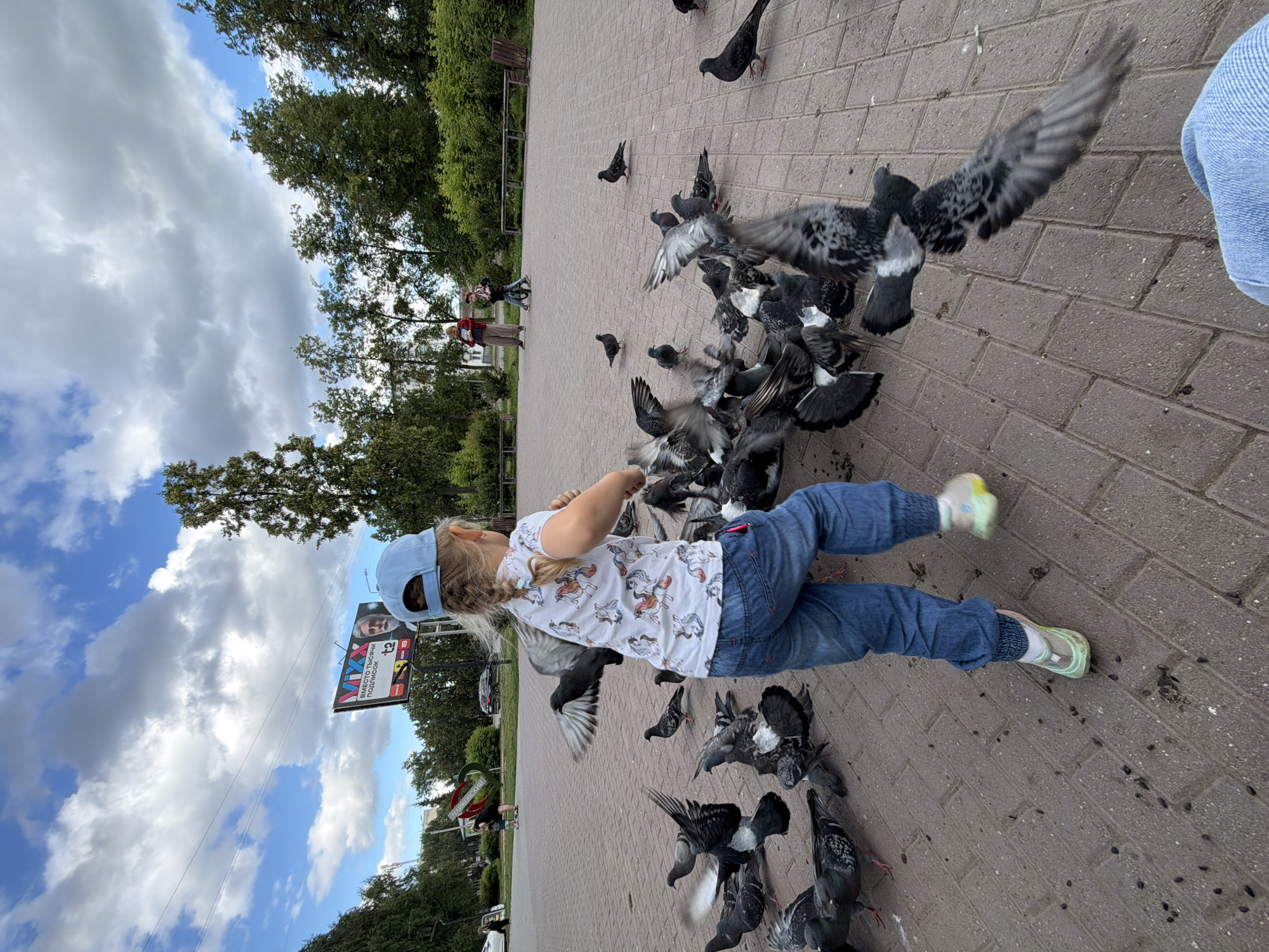 A young girl wearing a blue cap, a white t-shirt with a unicorn print, and blue jeans is the primary subject in this photograph, taken outdoors in Chaikovsky, Russia. She is interacting with a large flock of pigeons on a brick-paved ground, with trees and a cloudy sky visible in the background. The girl appears to be feeding or playing with the birds, with her arms extended towards them. Many pigeons are gathered around her, some in flight, while others are on the ground.
In the background, two adults are visible, one wearing a red shirt and the other in a beige outfit, also seemingly interacting with pigeons. Further in the distance, there are more trees and some buildings, suggesting an urban park or public space. A billboard with Russian text is also visible in the background, near the trees and sky. The weather appears to be overcast with some patches of blue sky, indicating it might be a partially cloudy day.
The overall scene depicts a candid moment of a child enjoying interaction with nature. The focus is on the girl and the pigeons, creating a lively and dynamic composition. The image seems to capture a moment during the day, likely daytime, given the lighting conditions. The presence of trees and the brick pavement suggest a park or walkway area in a town or city.