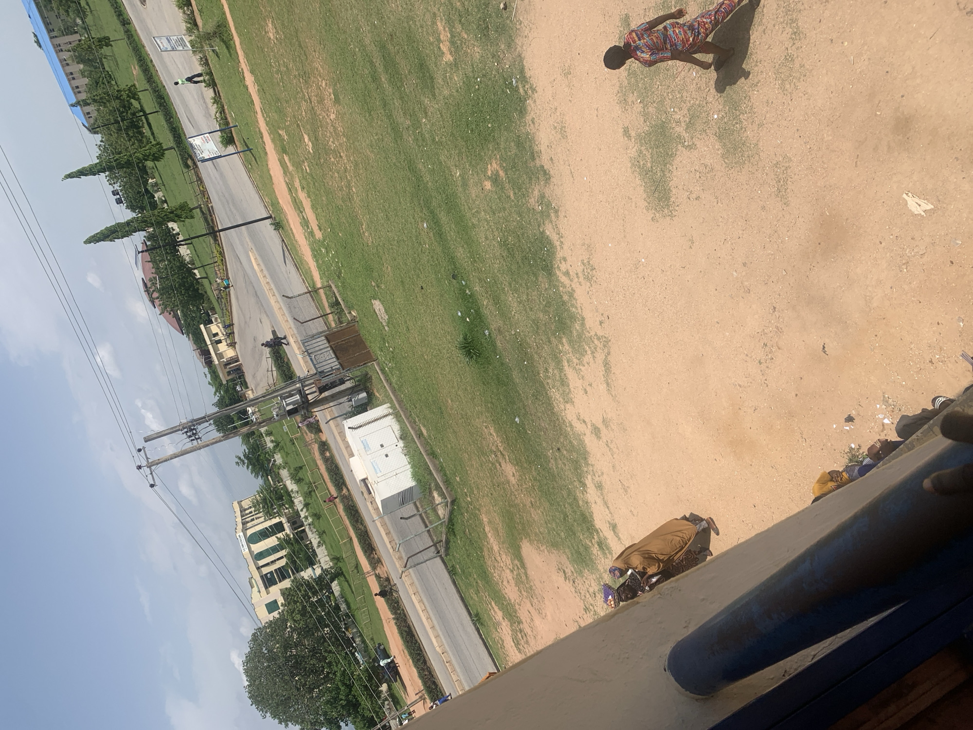 The image shows an outdoor scene with several people and buildings. The perspective is from a high vantage point looking down and to the side. In the foreground, a person wearing a brown garment is kneeling on a sandy area. To the right, another person in a colorful, patterned outfit is standing on the same sandy ground.
Further into the scene, a paved road runs alongside a grassy area. Several trees are planted along the side of the road. Buildings are visible in the background, including a multi-story structure with visible windows. Electrical poles and wires are also present, crossing the scene. The sky is mostly clear with some clouds, suggesting daylight. The overall setting appears to be a public space, possibly a campus or park, in Jalingo, Nigeria.
One building has text that appears to read "INDEPENDENCE HALL" in English. The activity appears to be casual, with people engaged in their own activities. The time of day is likely daytime given the lighting.