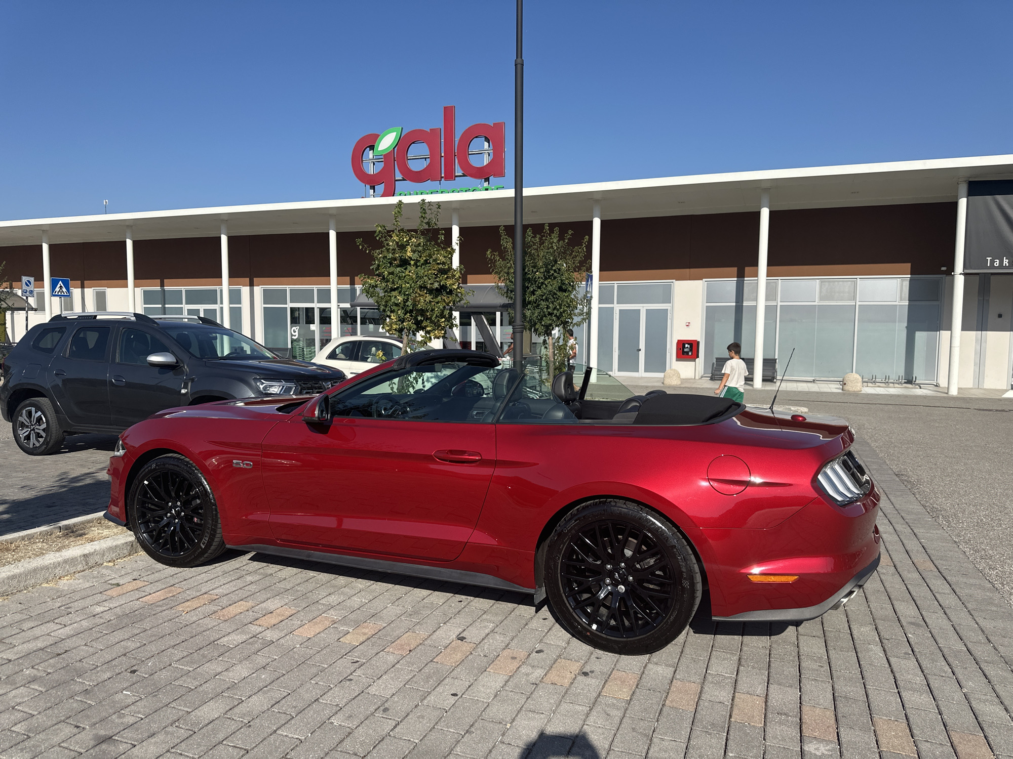 A red convertible Ford Mustang with black rims is parked on a brick-paved surface in front of a building with a large "gala" sign. To the left of the Mustang, a dark gray SUV is parked. The building appears to be a commercial establishment, possibly a supermarket, with large windows and white pillars. A young boy in a white t-shirt and green shorts is walking away from the building. The sky is clear and blue, and the sun is shining. The location is in the city of Fermignano, Italy.