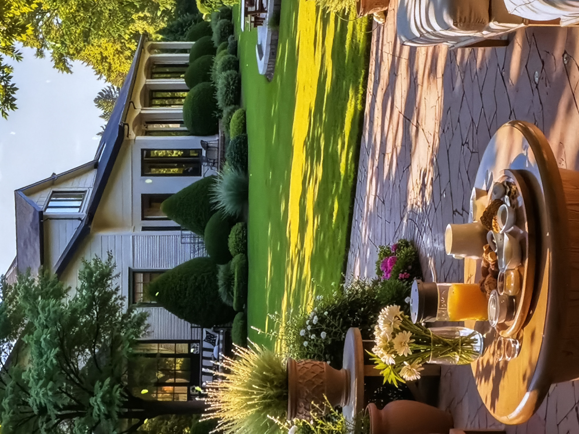 This image captures a picturesque outdoor scene of a residential property, likely a backyard in Sofiivska Borshchahivka, Ukraine. The main subject is a large, elegant house with a light-colored facade, a dark roof, and numerous windows, including a prominent gabled section and an upper-story porch or balcony.
The garden is immaculately maintained, featuring a vast, vibrant green lawn that stretches across the midground. This lawn is bordered by meticulously sculpted hedges in various spherical and conical topiary shapes, creating a refined landscape. Other lush green plants and bushes, some adorned with small pink and purple flowers, add to the verdant setting. Large, leafy trees with bright green foliage frame the scene, casting long, distinct shadows across the sun-drenched lawn and the adjacent paved patio area.
In the foreground, a round wooden outdoor table is set with items suggesting a relaxed al fresco meal or refreshment. Visible objects on the table include a clear vase holding a bouquet of fresh white daisies, a clear bottle or shaker filled with orange liquid (likely juice), and a wooden tray presenting several small bowls and cups containing snacks such as nuts or dried fruits, along with a white mug. Various potted plants, including one with spiky green foliage in a patterned terracotta pot, are also part of the foreground arrangement.
The strong, elongated shadows and the warm, golden sunlight illuminating the lawn and parts of the house indicate that the photograph was taken either during the early morning or late afternoon. The scene conveys a sense of tranquility, luxury, and peaceful outdoor living. There are no people visible in the image, nor is there any discernible text.