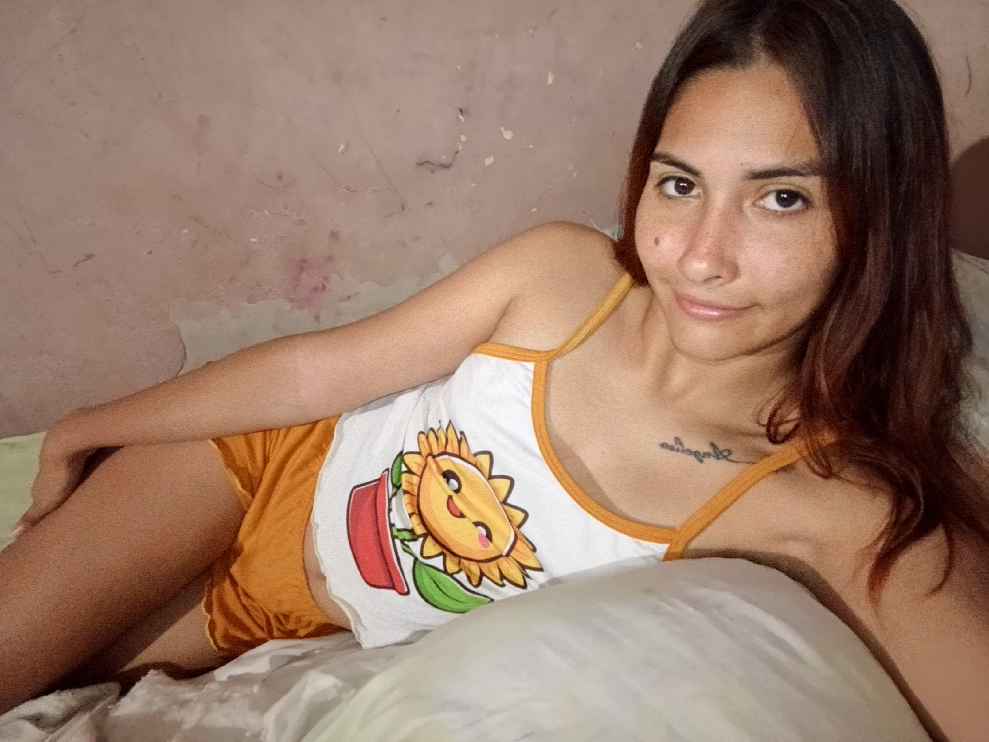 A young woman with dark hair and brown eyes is lying on a bed. She is wearing an orange camisole with a cartoon sunflower print and orange shorts. Her left arm is bent at the elbow, and her right arm is resting on a pillow. Her body is angled towards the camera, and she is looking directly at the viewer. A tattoo is visible on her upper chest. The background appears to be a wall with peeling paint. The lighting suggests an indoor setting. There are no other people or objects of note in the frame. The scene does not contain any specific visual cues that would indicate a location in Juan Griego, Venezuela.