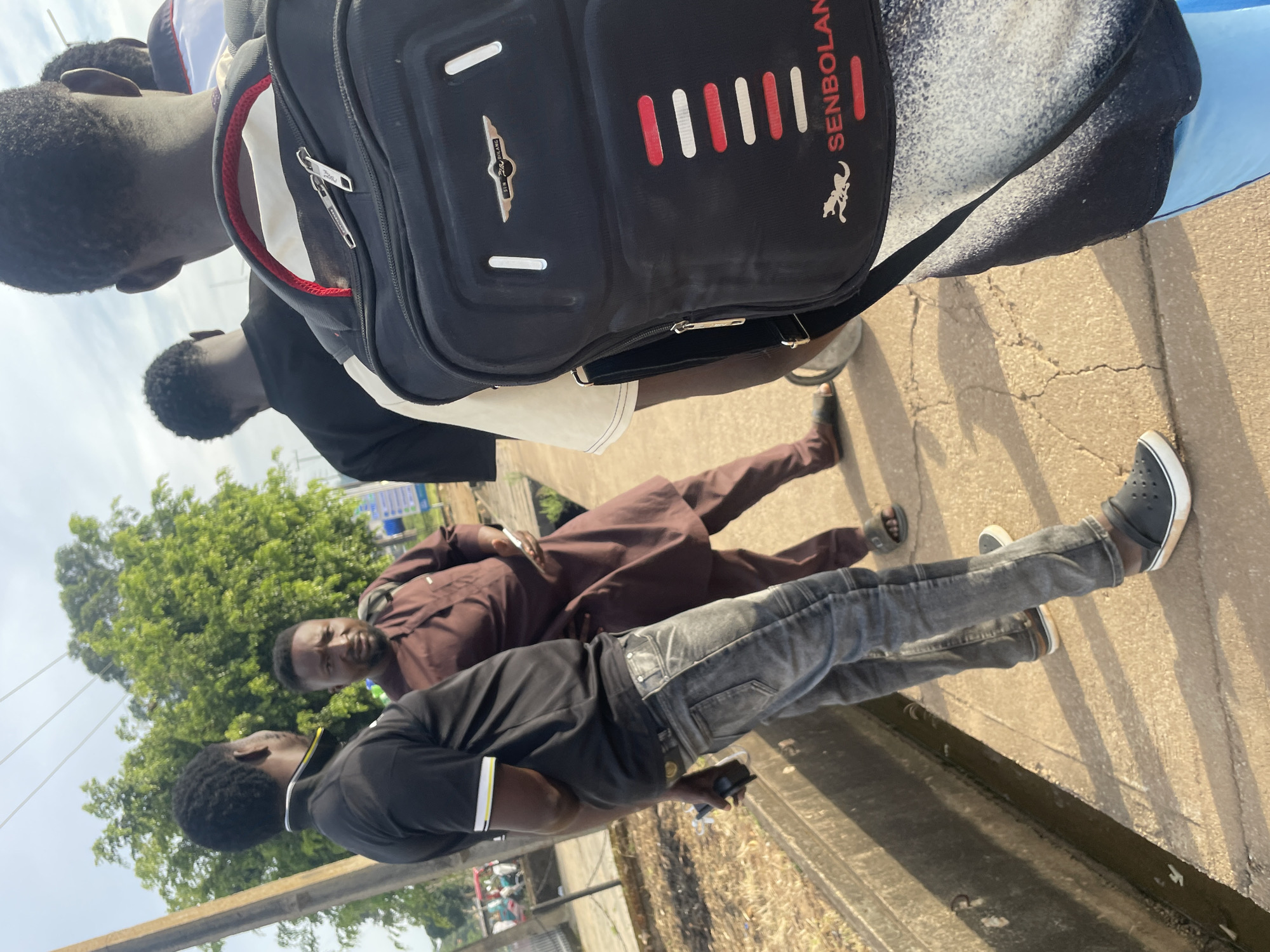 In Barakin Lerawi, Nigeria, four individuals are standing on a paved walkway beside a drainage ditch. The foreground is dominated by the back of a person wearing a black and white patterned t-shirt and light blue shorts, with a black backpack. To the left, a man in a black polo shirt and dark gray jeans stands facing away from the camera, holding a phone. To his right, another man in a maroon traditional outfit is speaking, holding a phone in his right hand. Further back, a fourth person, mostly obscured, is seen from behind, wearing a black shirt. The environment features a large green tree and a cloudy sky, suggesting it is daytime. The overall scene depicts a casual outdoor gathering on a street.
