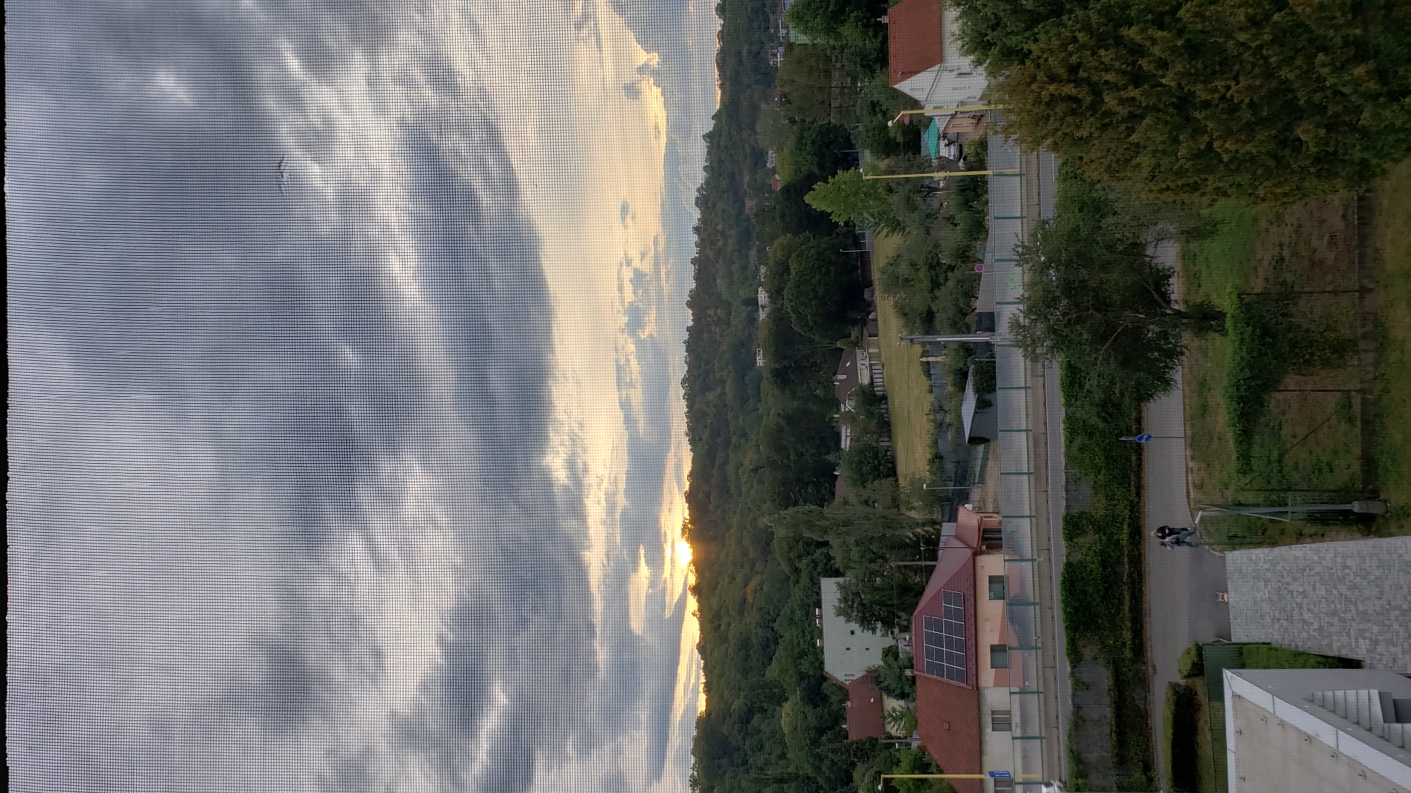 This image captures a dusky view of a residential area in Prešov, Slovakia, as seen through a window screen. The sky is dominated by heavy, grey clouds, with a hint of the setting sun breaking through on the horizon, casting a warm glow over the tree-covered hills. In the middle ground, several houses are visible, some with red roofs, and one with solar panels. A road runs horizontally across the image, with two individuals walking on the sidewalk. The foreground shows the edge of a modern building and a paved pathway. The overall scene suggests a quiet evening in a suburban neighborhood.