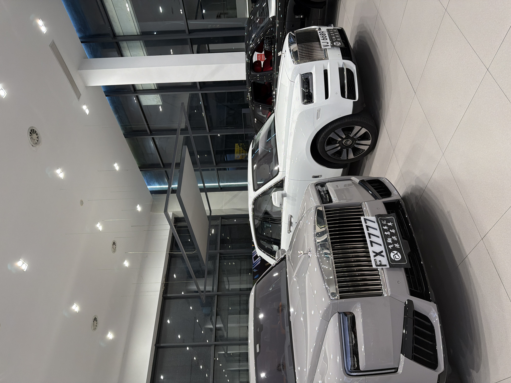 The image depicts a showroom displaying three Rolls-Royce vehicles in the city of Dongguan, China. In the foreground, a light grey Rolls-Royce features a prominent grille and a license plate reading "FX 7777". To its right, a white Rolls-Royce is visible, also with a distinctive grille and a license plate "VJ 6689". Behind these two, a black Rolls-Royce with a red interior can be seen. The showroom has a modern design with a white ceiling, recessed lighting, and large windows that reflect the interior. The floor is tiled in a light color. There are no people visible in the image.