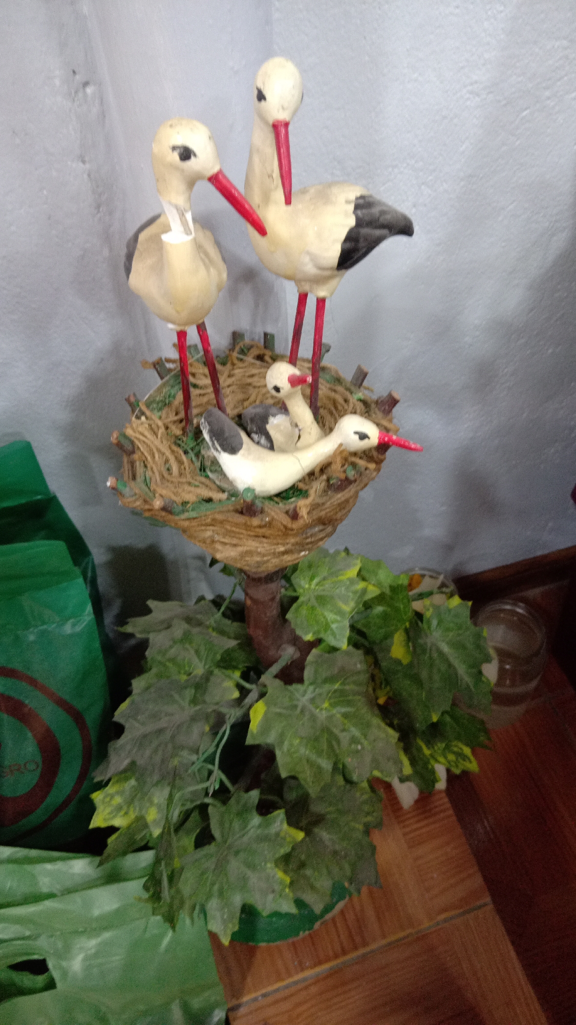 The image shows a decorative stork nest display. Two adult storks with red beaks and legs stand behind a nest. Inside the nest are two baby storks. The nest is made of brown twine and some green material. The display is set on a base of artificial green leaves, resembling a plant. A green plastic bag is visible on the left, and a glass jar on the right. The background is a plain, light-colored wall. The location, Yerevan, Armenia is based solely on the user request.