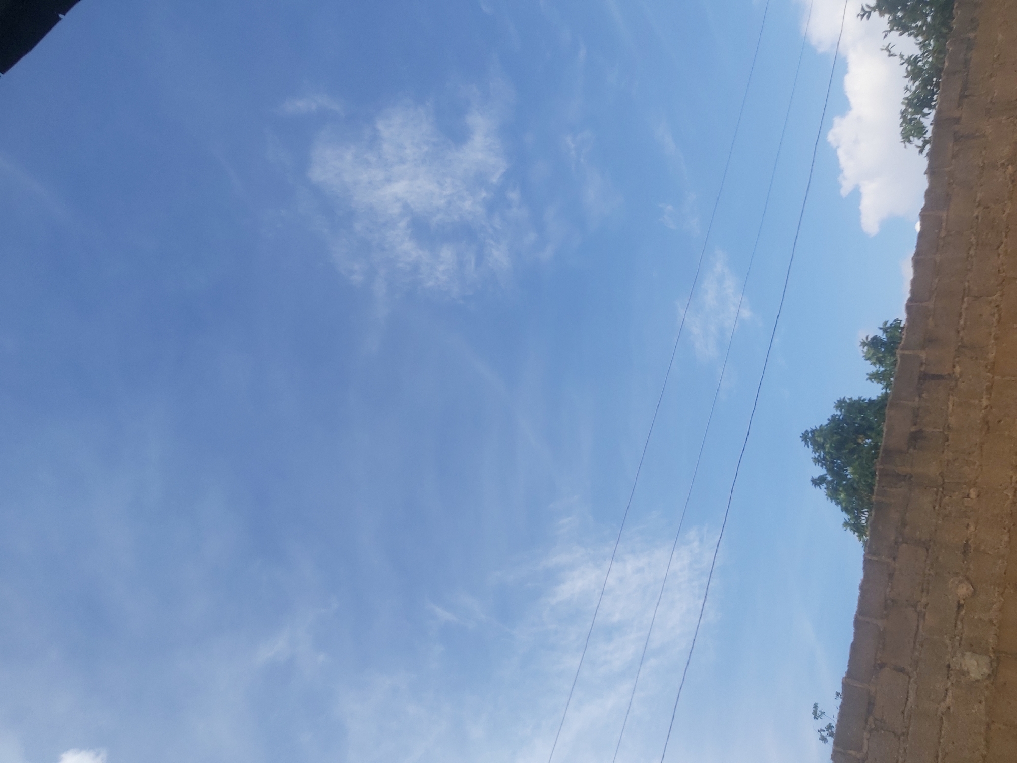 The image depicts a clear blue sky with scattered wispy clouds, characteristic of a sunny day. In the foreground, a rough-textured, light brown brick wall runs horizontally across the bottom of the frame. Above the wall, the tops of several trees with green foliage are visible, suggesting an outdoor setting within a populated area, possibly a residential or commercial district. Several power lines are strung across the sky, indicating the presence of infrastructure. The overall scene suggests a typical urban or suburban environment in Jos, Nigeria, on a bright day.