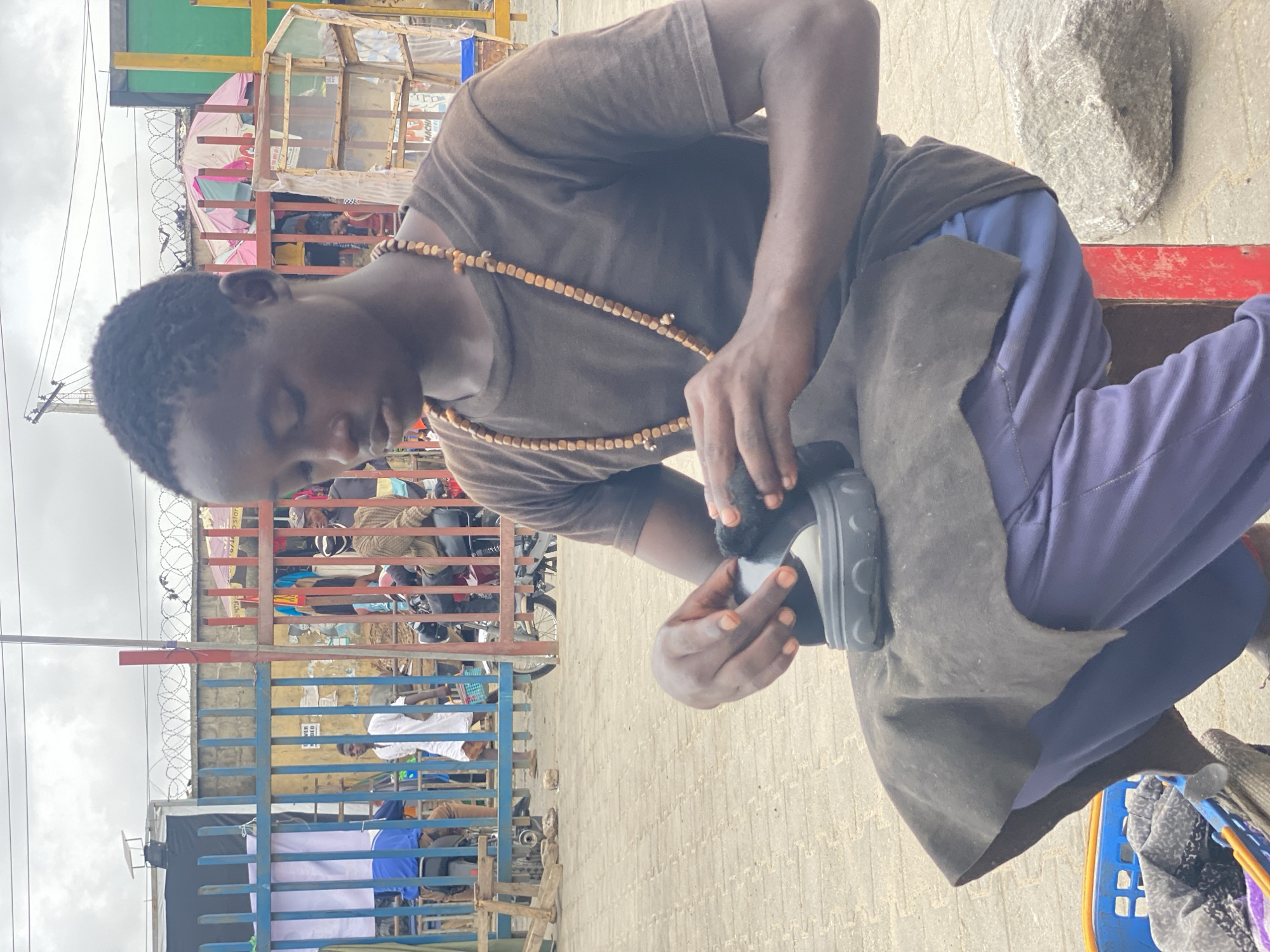 A young Black man is depicted in an outdoor market setting, intently engaged in polishing or repairing a shoe. He wears a brown short-sleeved t-shirt, blue sweatpants, and a distinctive long necklace made of light-brown beads. A dark gray piece of fabric covers his lap, likely functioning as an apron or protective cloth. He holds a dark-colored shoe in one hand and a small, dark brush or applicator in the other, focusing on his task.
The background reveals a vibrant and informal market environment. Numerous makeshift stalls and structures, constructed from materials like wood, corrugated metal sheets, and tarpaulins, are visible. A blue metal fence, topped with barbed wire, spans across the mid-ground, separating the immediate area from further market activity. Other individuals can be glimpsed beyond the fence. The ground is paved with light-colored interlocking bricks. To the right, a large, gray, irregular rock sits on the ground, and a blue plastic basket containing various items is partially visible in the bottom right corner.
The scene occurs during the daytime under an overcast sky, providing soft, even lighting. The man appears focused and diligent in his work. While some text is faintly visible on background signs, it is too obscured to be legibly identified. The setting suggests a bustling urban market, consistent with locations in Lagos, Nigeria.