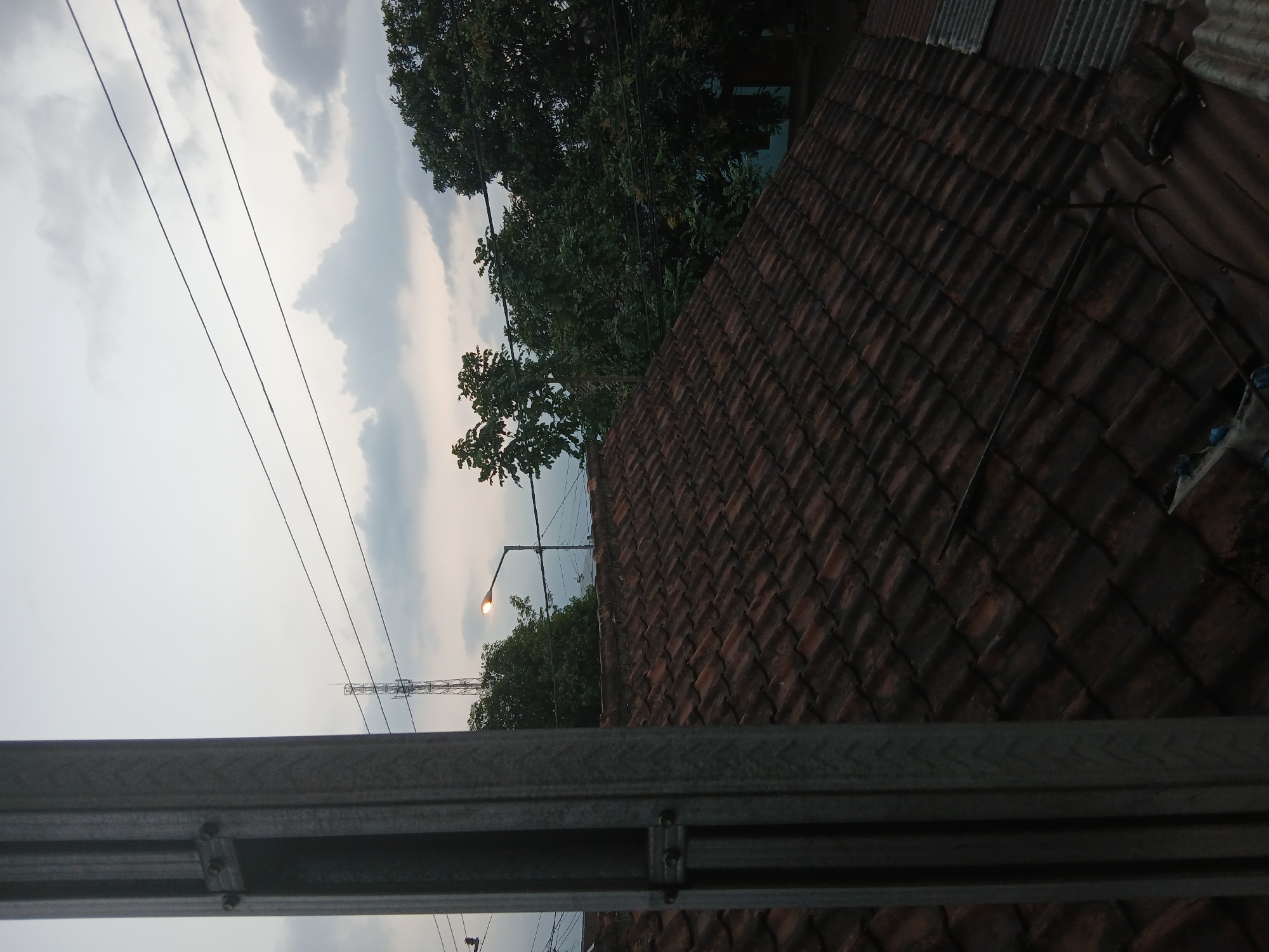 The image depicts an outdoor scene from a high vantage point, likely a rooftop, in Kademangan Kulon, Indonesia. The foreground is dominated by a large, gray metal support structure on the left, which appears to be part of a building's framework. To the right and extending into the midground is a steeply sloped roof covered with weathered, reddish-brown terracotta tiles. Beyond the tiled roof, lush green trees fill the midground and background, with dense foliage characteristic of tropical vegetation. Above the trees, a utility pole with a single, illuminated streetlamp is visible. Further in the distance, a tall telecommunications tower stands against a cloudy sky. Power lines crisscross the sky, connecting various unseen structures. The overall ambiance suggests it is either early morning or late afternoon, with the sky a muted gray due to cloud cover. No people or animals are present in the frame.