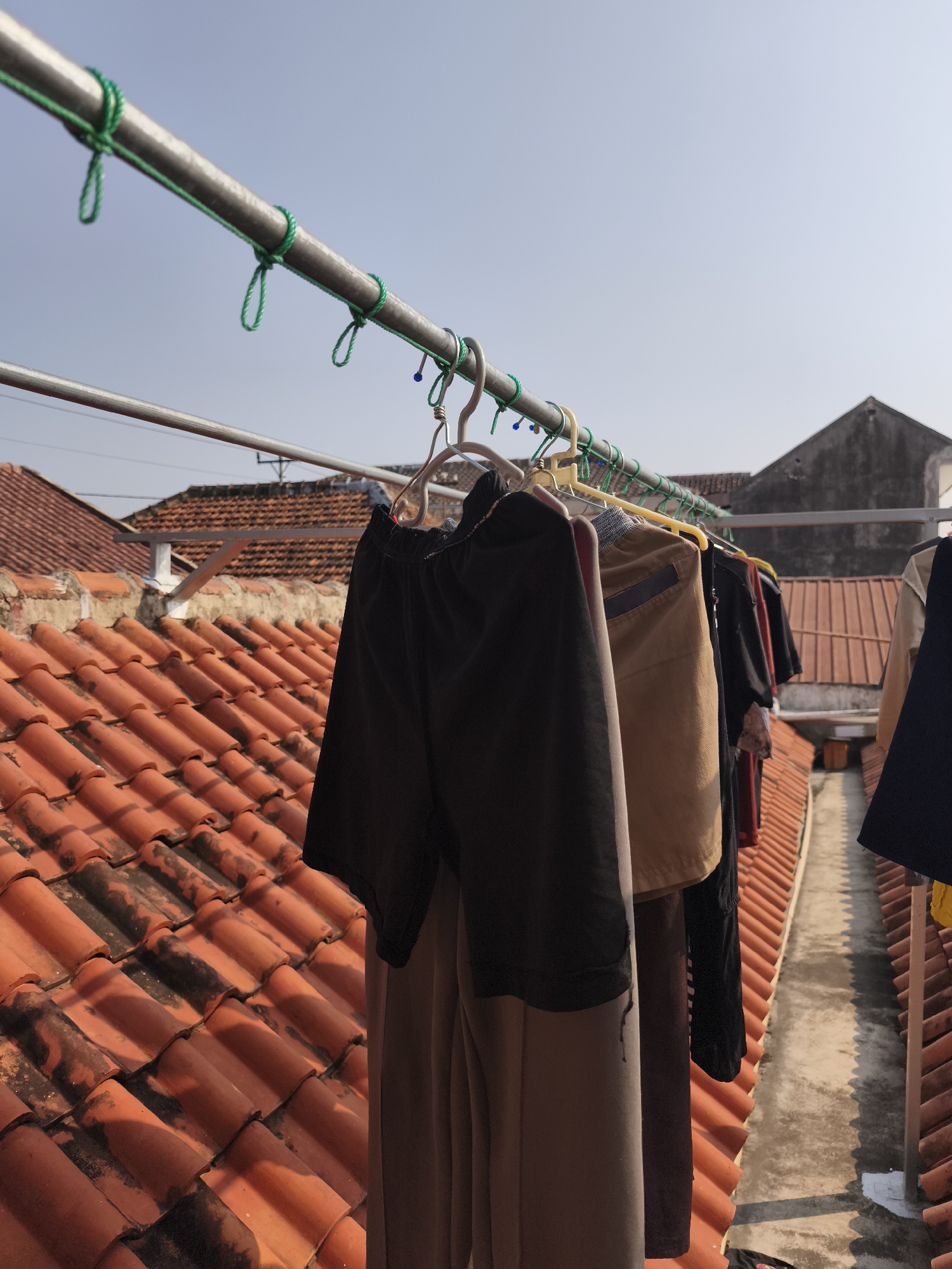 The image shows clothing hanging on a clothesline outdoors. The clothesline is a metal pipe strung with green rope, attached to a larger metal structure above. Several items of clothing, including shorts and t-shirts in various colors, are hanging on hangers. In the foreground, the textured, orange-red tiles of a rooftop are visible, casting shadows. Behind the clothesline and rooftop, the grey, sloped roofs of residential buildings are visible, along with a clear blue sky. The scene appears to be in a residential area in Yogyakarta, Indonesia, suggested by the characteristic red tile roofs and the overall urban residential aesthetic. The lighting indicates it is daytime.