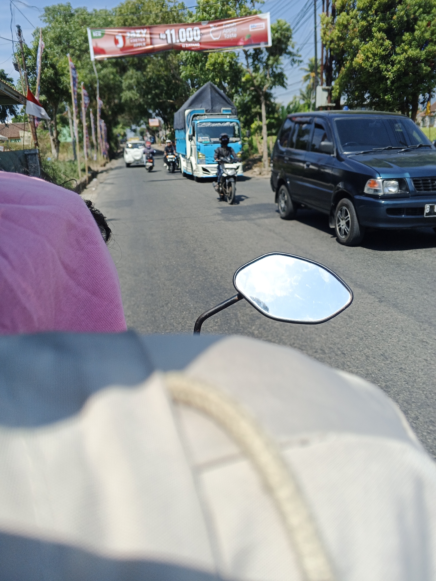 The image captures a street scene in Sukabumi, Indonesia, likely taken from the perspective of a motorcycle passenger. In the foreground, a portion of a pink fabric and a blurred white fabric are visible. A motorcycle's side mirror, reflecting the sky and clouds, is also prominent. The road ahead is lined with trees and flags, including a banner with text that appears to be an advertisement, possibly for a product or event. Several vehicles are on the road: a blue and white truck, a dark-colored minivan, and multiple motorcycles with riders. The environment suggests a sunny day, with a clear blue sky visible above the trees and vehicles. The presence of a banner and flags indicates a public area within the city.