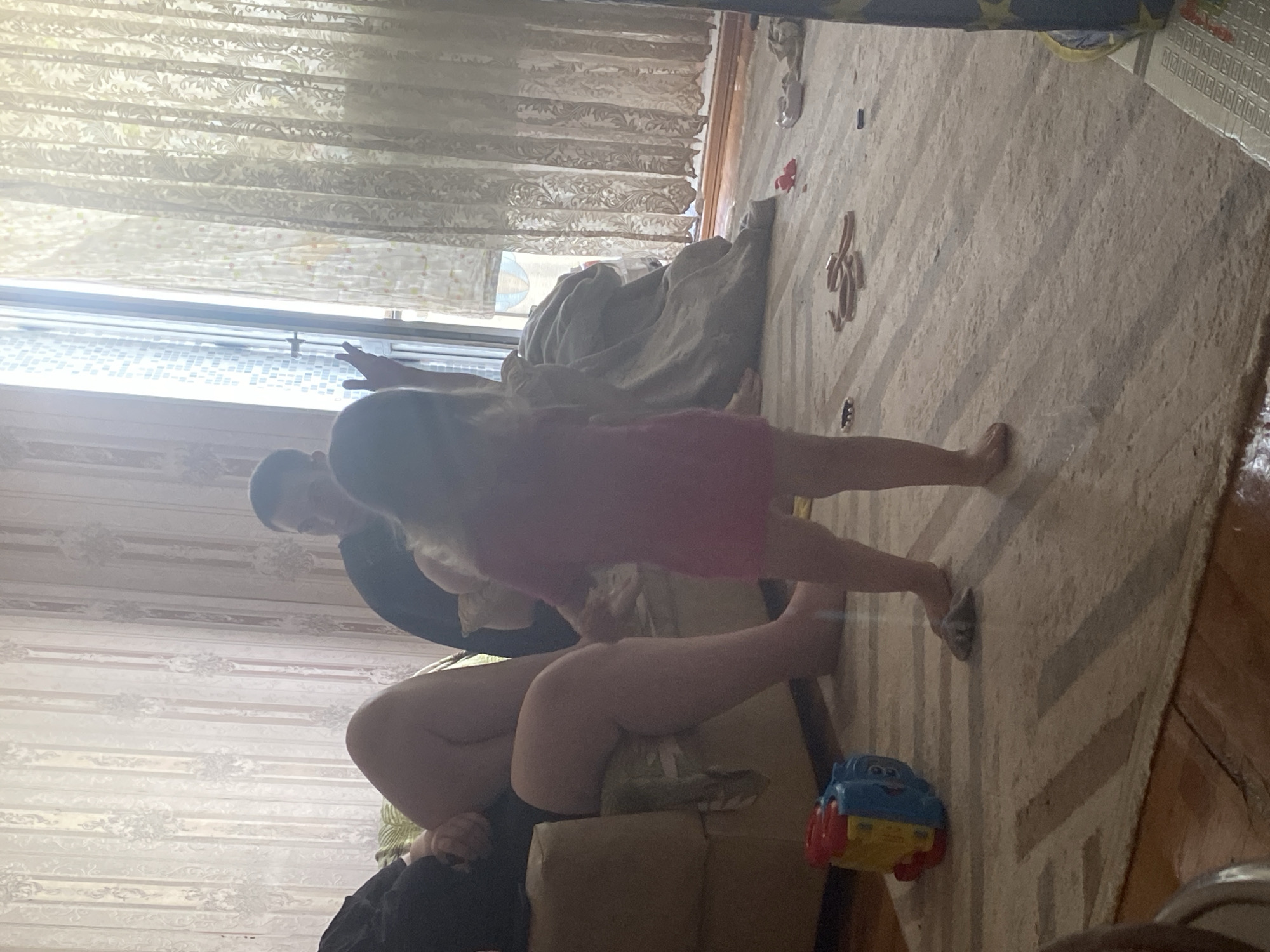 The image depicts an indoor scene, possibly a living room. A young child, wearing a pink dress, stands in the center of the frame, facing away from the camera. To the left, a person is seated on a sofa, their legs visible. In the background, another person is partially visible. The room is furnished with a sofa and a rug on the floor. Toys, including a multi-colored toy, are scattered on the rug. The scene is illuminated by natural light streaming through a window with curtains. The overall impression is of a casual, domestic setting. The image does not contain any specific visual cues that would allow for precise localization within the city of Shakhtinsk, Kazakhstan.