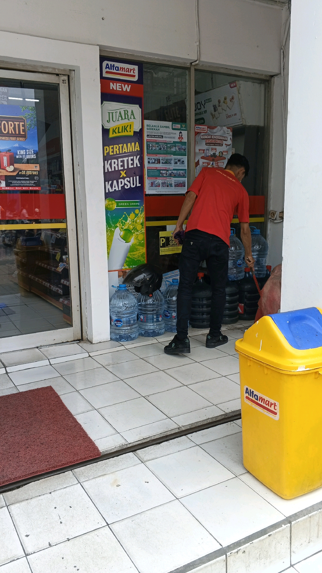 A man in a red shirt and black pants is standing outside an Alfamart convenience store in Balaraja, Indonesia. He is holding a broom and appears to be sweeping the tiled floor. In front of the store are several large bottles of water, a black helmet, and a yellow trash bin with the Alfamart logo. The store entrance has glass doors and windows displaying various advertisements and products.