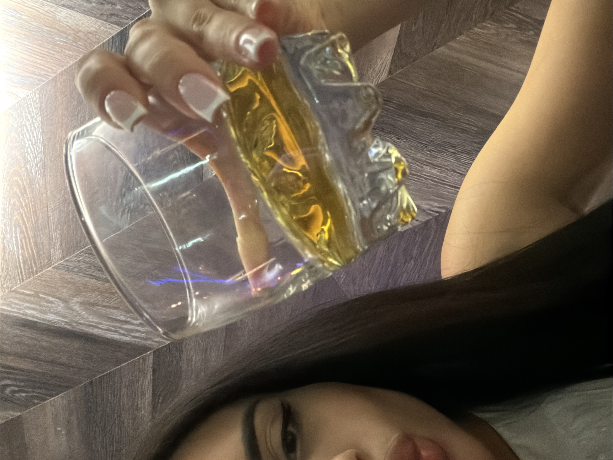 A low-angle shot shows a woman with long, dark hair and fair skin. Her right eye and part of her face are visible. She is holding a glass containing a golden liquid, possibly whiskey or juice. Her hand is visible, with manicured nails featuring a white French tip. The background consists of a surface with a wood-like texture. The scene is indoors. There is no evidence to suggest this photo was taken in Burgas, Bulgaria.