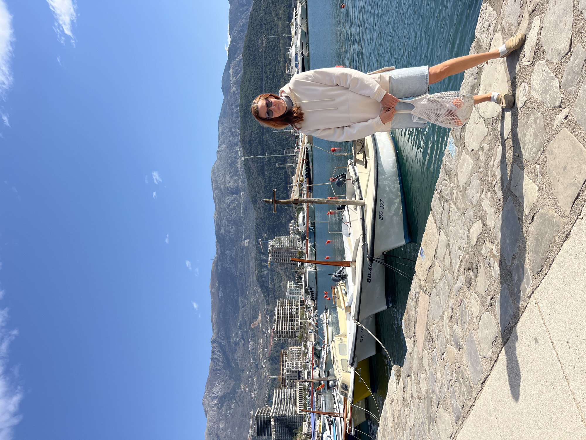 A woman stands on a stone pier in Budva, Montenegro, with boats docked to her right and mountains in the background. The woman, wearing sunglasses, a cream-colored hoodie, light denim shorts, white socks, and tan slip-on shoes, is holding a white mesh bag. Several boats are visible, including one with the designation "BD 817" and another with "BD-44." The city buildings are visible in the background. The sky is blue with scattered clouds. The pier is constructed of irregular stone blocks with a smooth, concrete edging.