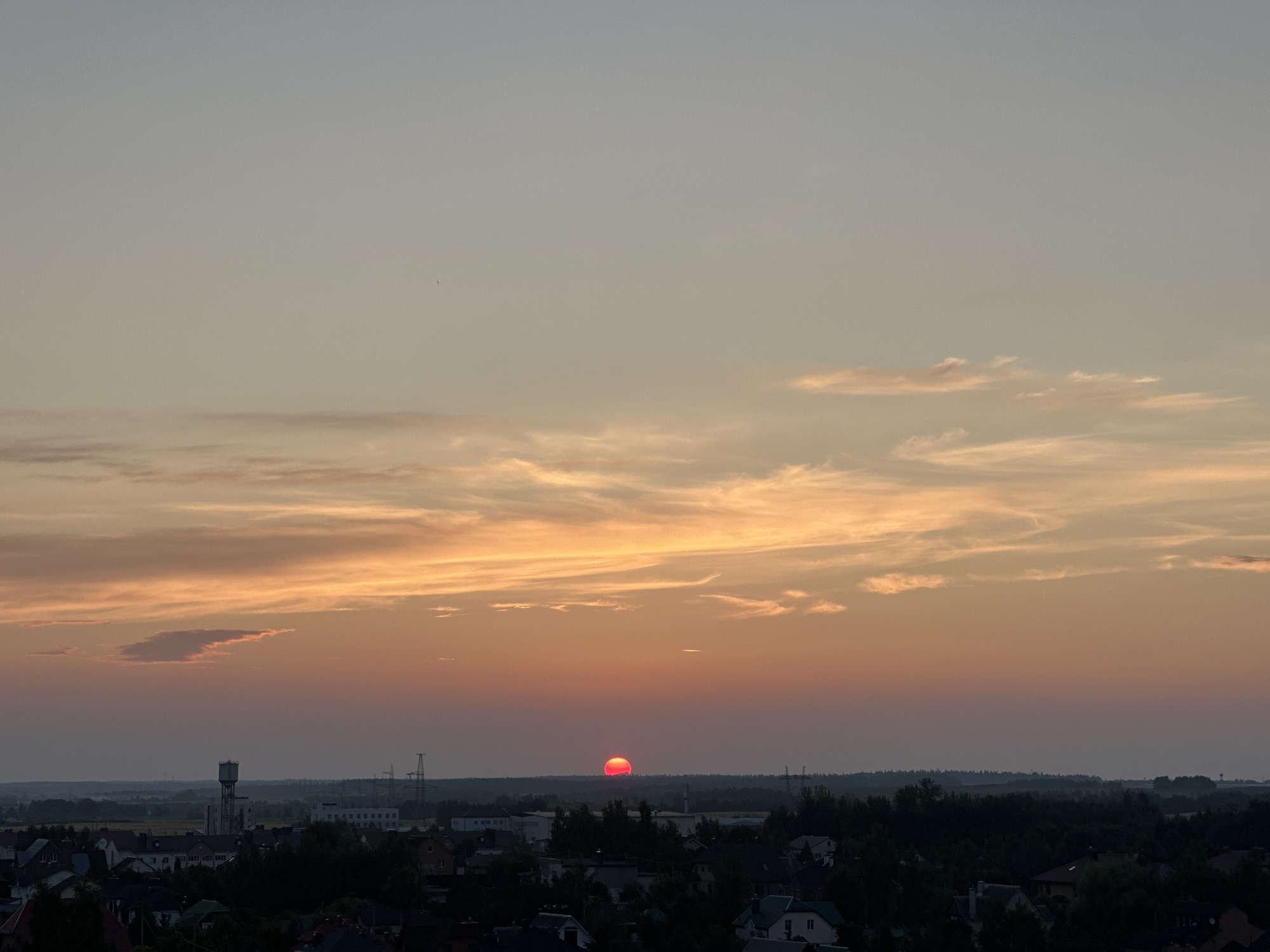 The image captures a sunset over a suburban landscape. The sky transitions from a pale blue at the top to warm hues of orange and pink near the horizon. Scattered clouds, illuminated by the setting sun, are visible. In the foreground, a dark silhouette of trees and rooftops of houses is present, suggesting a residential area. A prominent water tower stands tall among the buildings, and power lines and pylons are visible in the distance, indicating infrastructure. The sun, a vivid red orb, is partially obscured by the horizon line, casting a warm glow over the scene. The overall impression is a peaceful evening in a rural or semi-rural setting, possibly within the Шчомысліцкі сельскі Савет, Belarus, given the context.
