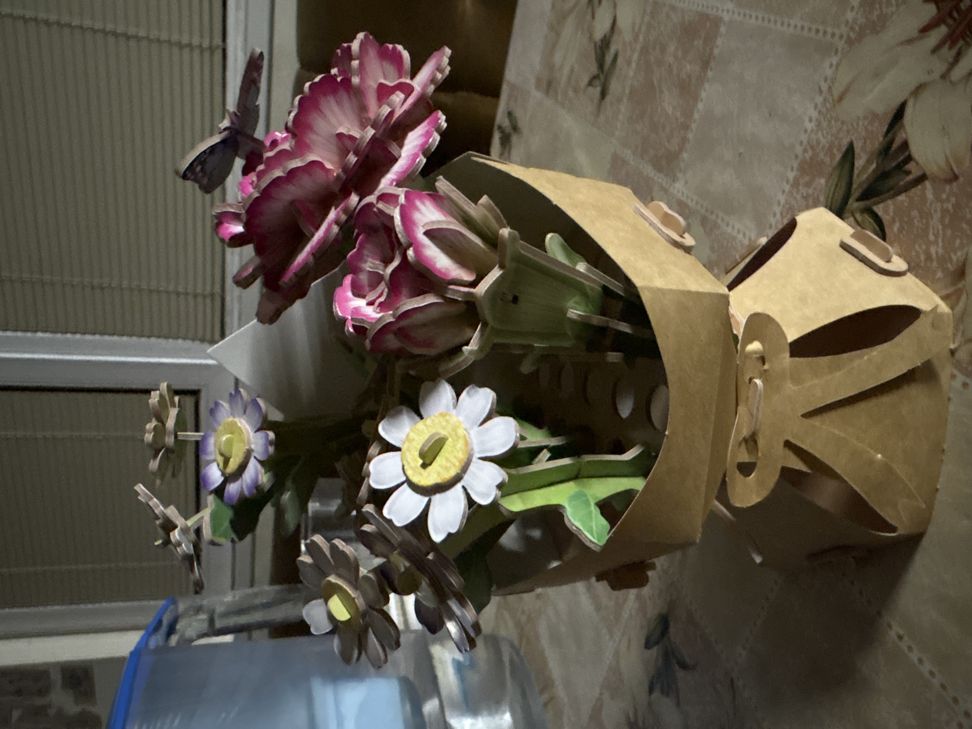 A close-up shows a bouquet of assembled cardboard flowers in a cardboard vase. The flowers include pink and white carnations, purple and yellow daisies, and white and yellow daisies, with a cardboard butterfly on one of the carnations. The vase is made of light brown cardboard with circular cutouts. The bouquet is on a table with a patterned tablecloth, and a blue object is visible in the lower left corner. The background is out of focus and shows a wall with blinds. The location is not discernible from this image.