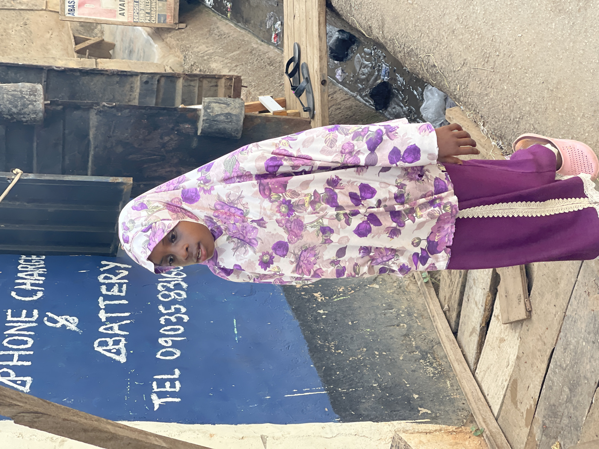 A young girl stands outdoors in a daytime setting in Kuchingoro, Nigeria, looking directly at the camera with a neutral expression. She is dressed in a white top with vibrant purple floral patterns, a matching head covering, purple pants adorned with white lace trim at the ankles, and light pink slip-on shoes.
To her left, a blue wall serves as a backdrop, featuring hand-painted white text that reads "PHONE CHARGE & BATTERY" and "TEL 0903583160," indicating a service available at this location. To her right and in the background, there is a concrete path running alongside an open drainage ditch. The ditch contains murky water and various pieces of debris, including what appear to be plastic bags. A pair of black flip-flops is seen hanging on a wooden post situated near the drainage area. Further in the background, a collection of wooden planks and structures is visible, along with a partial sign at the upper right. This sign shows fragments of text such as "ABAS," "AVAI," "DEPOSIT," "SEND MOR," "PAY BILLS," and "CHARGING," suggesting financial or telecommunication services. The bright, even lighting indicates that the photograph was taken during the day in an urban or semi-urban environment.