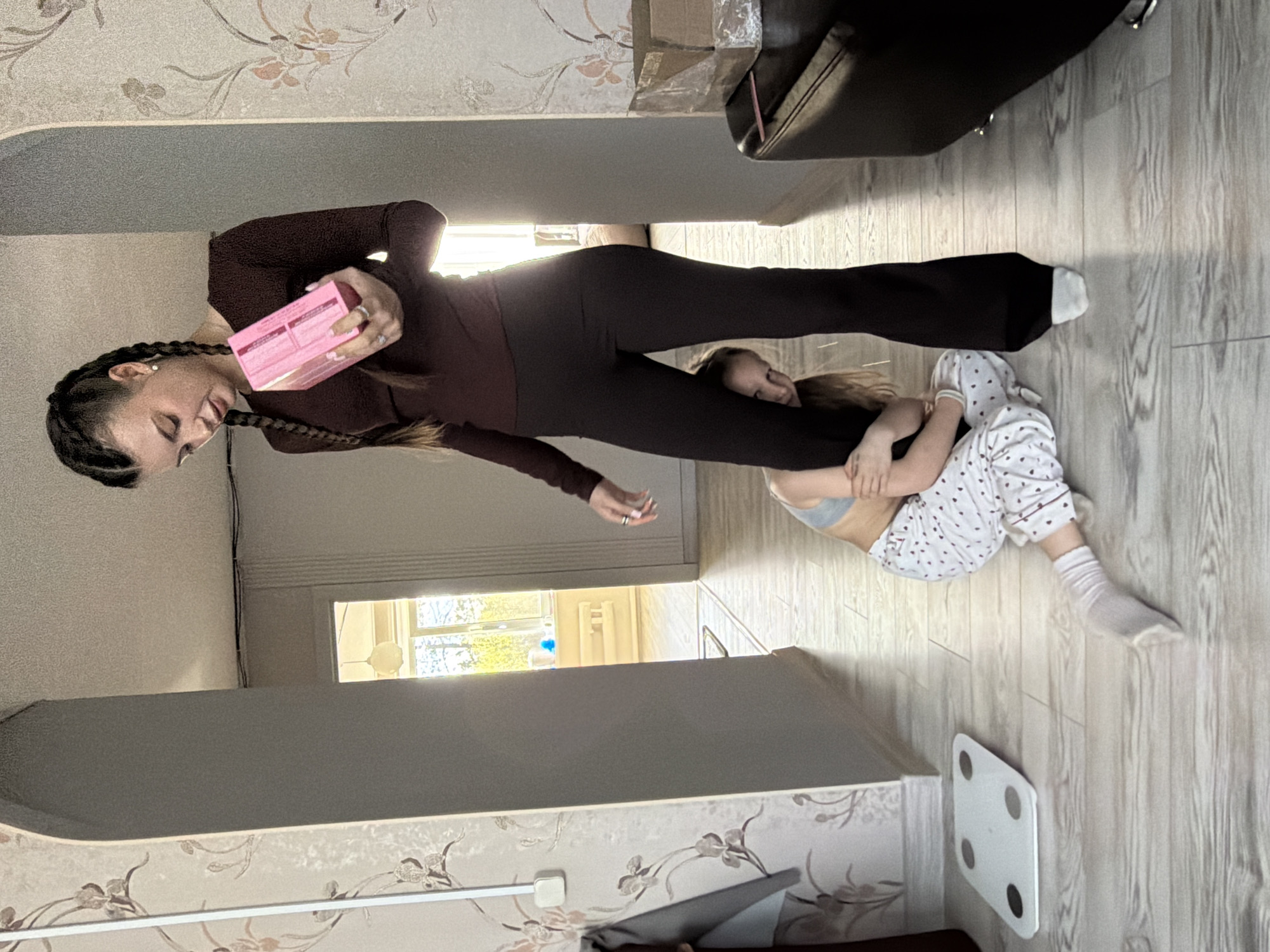 A woman stands over a child lying on the floor of a domestic interior in Dubovaya, Russia. The woman wears a maroon long-sleeved top and leggings and holds a pink rectangular object. Her hair is styled in braids. The child is dressed in white pajamas with a heart pattern and white socks, embracing the woman's legs. The room features floral wallpaper, light-colored wood flooring, and a digital scale. A black sofa is visible in the background. The image suggests a playful or affectionate interaction.