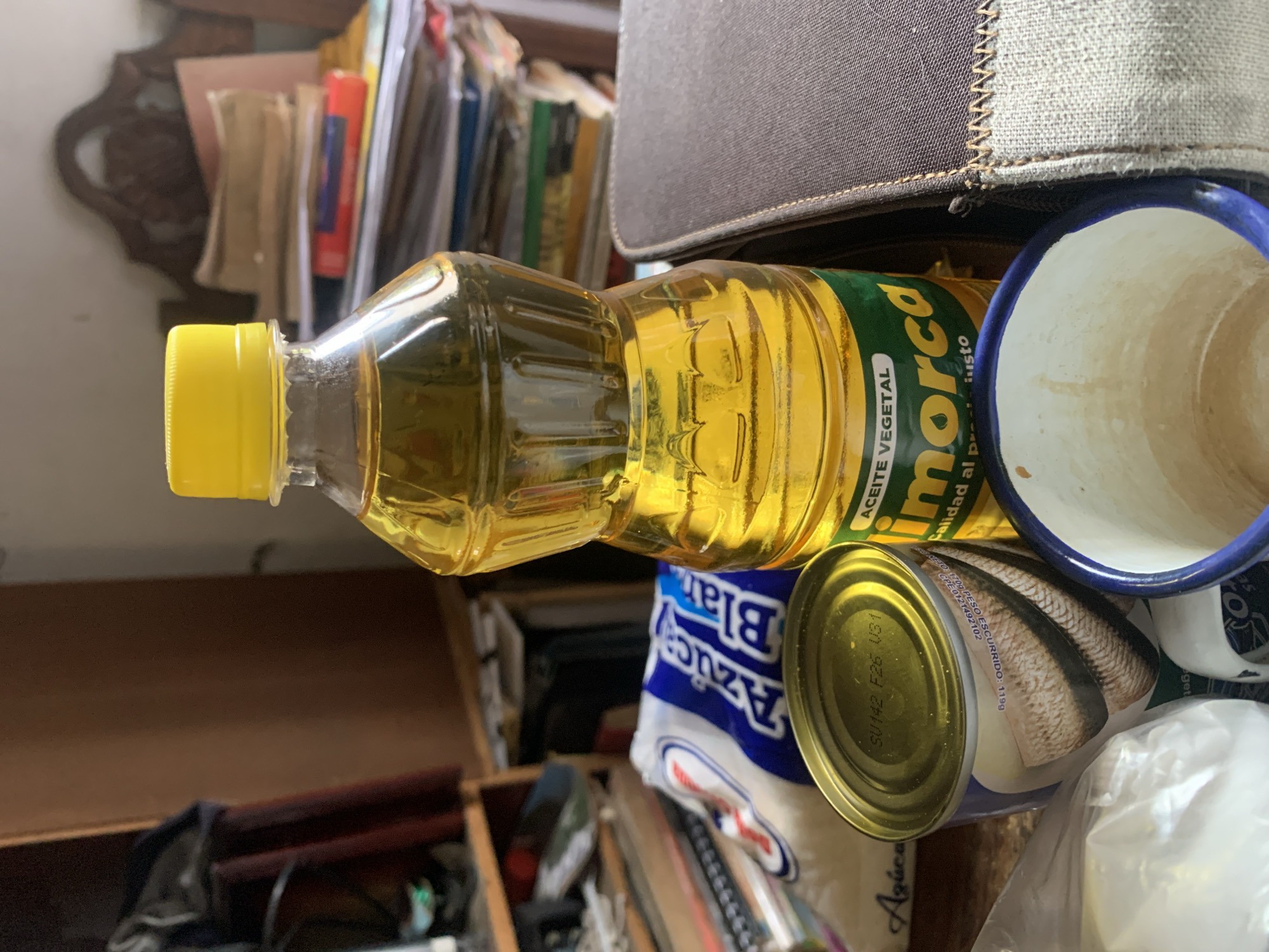 This image captures a still life of various household items, likely on a shelf or surface, set in Santa Ana, Venezuela. The scene is shot at an angle, making the objects appear tilted.
The central subject is a clear plastic bottle filled with yellow cooking oil, featuring a bright yellow cap. Its label, partly visible, reads "morca", "ACEITE VEGETAL", and "Calidad al gusto". To the right of the oil bottle, a white and blue ceramic mug is visible. Below the oil bottle, there's a metal can with a gold-colored lid. The can's label shows an illustration of fish and includes text like "NETO 170g PESO ESCURRIDO: 119g", "CPE 0121492102", and "SU142 F26 U31". Partially hidden behind the can and oil bottle is a white bag with blue lettering, clearly showing "Azúca" and "Blar", indicating "Azúcar Blanco" (White Sugar). Above these items, a dark gray fabric object with stitching, possibly a bag or part of furniture, occupies the upper right portion of the frame.
The background is out of focus, revealing a cluttered space with numerous books, papers, and what appears to be dark wooden decorative elements and shelving, suggesting an indoor setting. There is no specific activity taking place, as the image is a static shot of stored goods. The lighting suggests ambient indoor conditions. The Spanish text on the product labels is consistent with the provided location context of Venezuela.