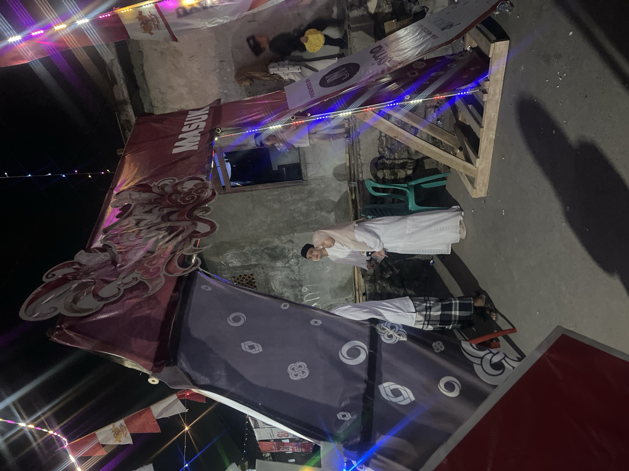 The image depicts an outdoor scene at night, likely in Klapanunggal, Indonesia, featuring an illuminated archway decorated with Indonesian flags and colorful lights. The archway is adorned with a large, dark purple banner on the left, featuring white circular and floral patterns. On the right, a red banner with white lettering that reads "MASUK" (meaning "Enter" in Indonesian) is visible. Below this, a triangular wooden structure supports a banner with text and a logo.
Several people are present near the archway. Two individuals in the foreground are dressed in traditional white clothing. One, a male, wears a black cap, and the other, a female, is in a headscarf. Further back, through the archway, a child is visible standing in an opening. To the right of the archway, other individuals, including children, are seen interacting. One child in the background is carrying a yellow object.
The environment appears to be a street or pathway in front of a rustic building with a rough concrete wall. Green plastic chairs are placed near the archway. The ground is paved and shows scattered debris. The overall atmosphere suggests a festive or celebratory event taking place in the evening.