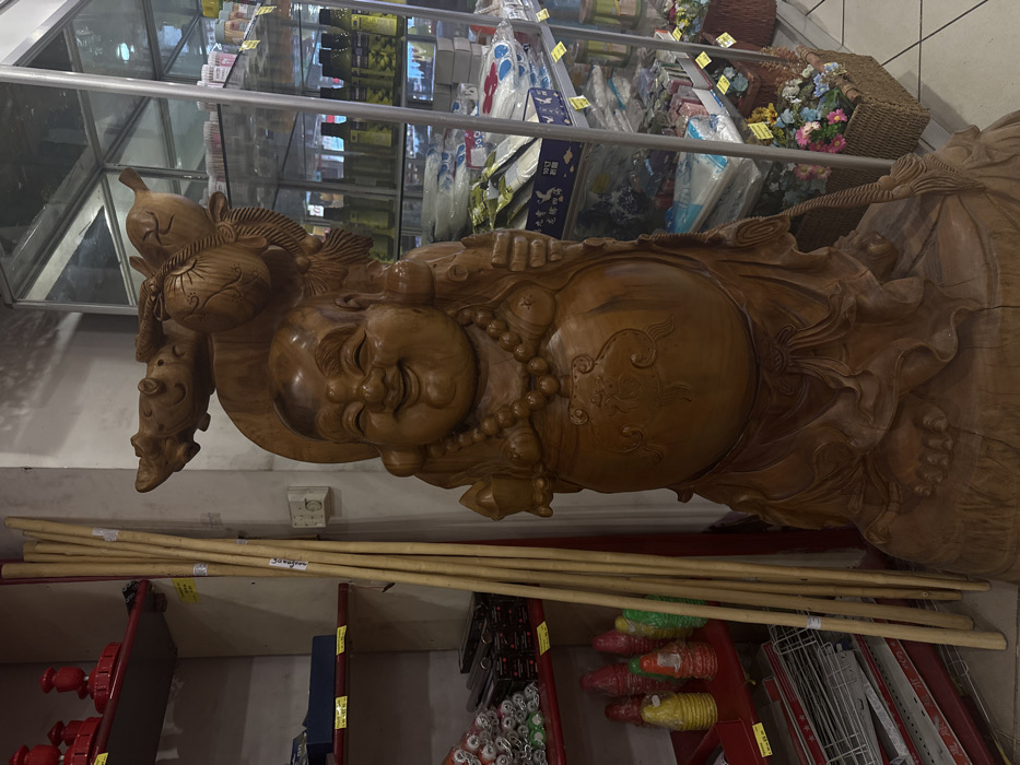 The image displays a large, intricately carved wooden statue of Hotei (the "Laughing Buddha") positioned in a retail environment, likely an Asian grocery store or market in Sofia, Bulgaria. The statue, rendered in light-toned wood, depicts a corpulent, smiling figure with a prominent belly, wearing a beaded necklace. It carries a large sack and a fish-like creature over its right shoulder and holds an object in its left hand. The figure stands upon a carved wooden base resembling a tree stump.
To the immediate left of the statue, a red metal shelving unit contains various goods: red ornamental objects on the top shelf, packaged items on a middle shelf, and stacks of yellow, red, and green cone-shaped plastic items on a lower shelf. Several long, light-colored wooden poles lean against a pale wall between the shelving unit and the statue. A white electrical outlet is visible on this wall.
To the right of the statue, a multi-tiered glass display cabinet is filled with various retail products, including bottles of liquids (possibly sauces or oils), packaged goods, and other merchandise. Yellow price tags are visible on the shelves. In the lower right corner, woven baskets containing artificial flowers are arranged on a white tiled floor. The scene is devoid of any individuals. While the type of establishment is identifiable as a specialty market within Sofia, the precise street address or specific name of the location cannot be determined from the image provided.