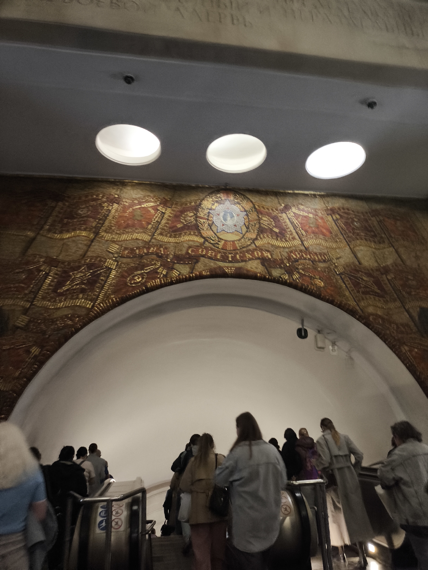 A wide-angle, medium shot taken indoors depicts a group of people riding up an escalator. The location appears to be a subway station, possibly in Unknown, Unknown. Overhead, a large, arched mosaic mural covers the wall, featuring red and gold colors and a central emblem with a star. Three circular light fixtures are embedded in the ceiling above the mural. The people on the escalator are diverse in their attire, and their backs are mostly visible. The environment is well-lit, with the light sources creating a bright atmosphere. The escalator itself has metal railings with instructional signs.