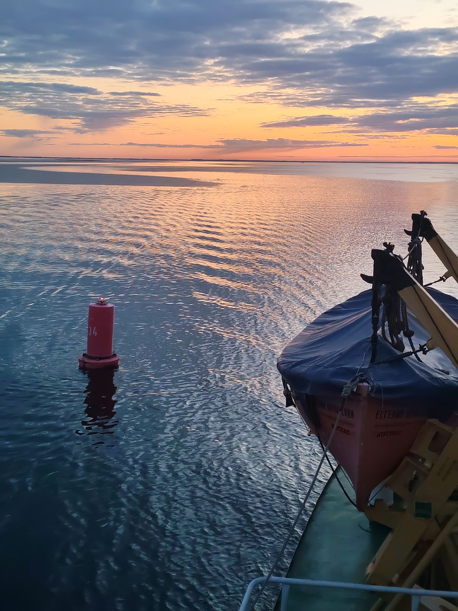 The image depicts a seascape at sunset, possibly in the vicinity of Kirgody, Russia. In the foreground, there is a lifeboat, secured to the side of a ship or vessel. A red navigational buoy, marked with the number 34, is visible in the water. The sea extends towards the horizon, reflecting the orange and pink hues of the sunset. The sky is partly cloudy, with shades of grey and blue intermingling with the sunset colors. The water shows gentle ripples, and the overall scene suggests a tranquil maritime environment.