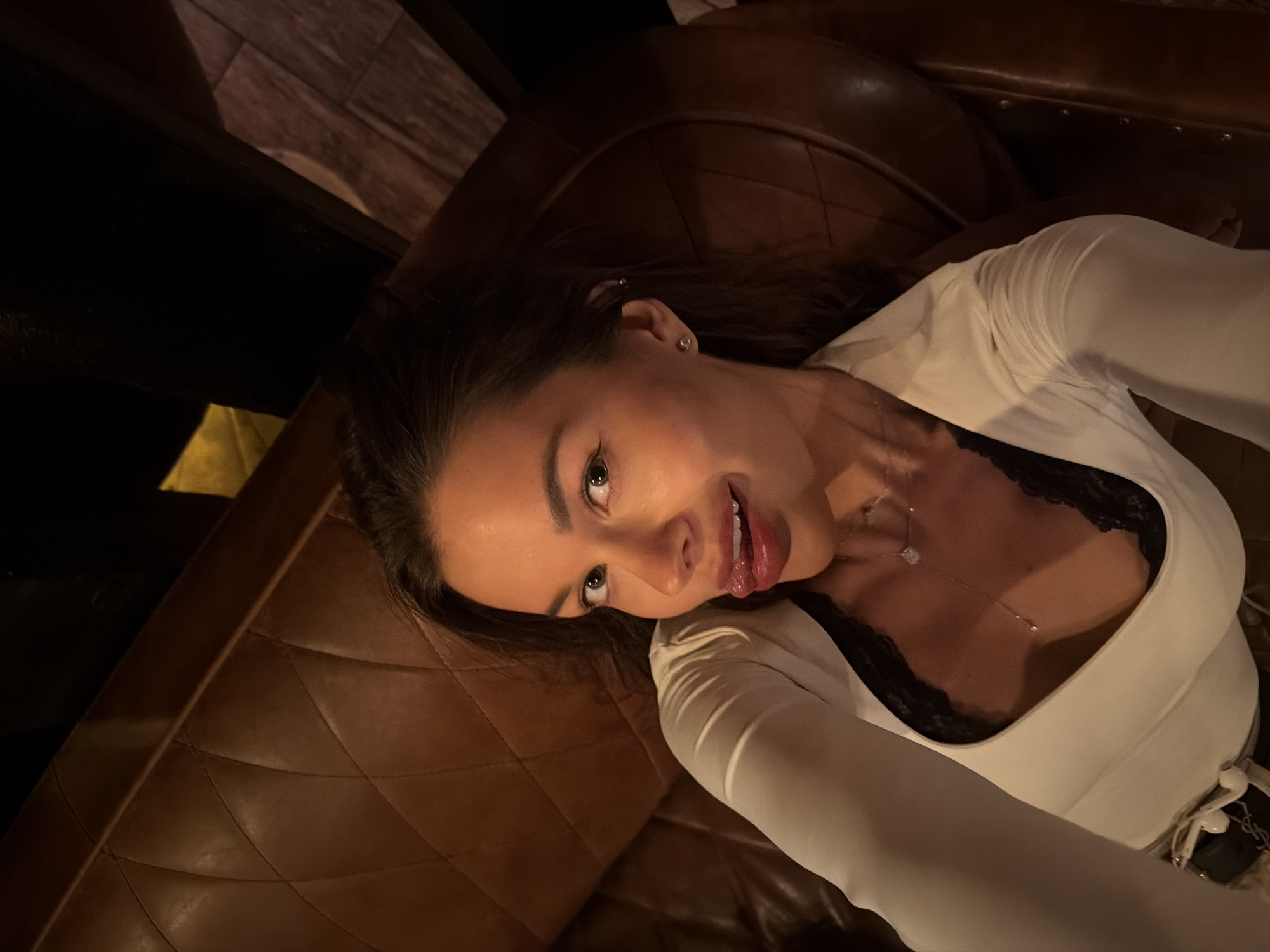 A young woman is captured in a selfie, lying or leaning back on a dark brown, quilted leather sofa or booth. Her head is tilted towards the upper right of the frame, and she looks directly at the camera with a playful expression, her tongue slightly out, and a hint of a smile. She has dark hair, styled down, and is wearing makeup including eyeliner and full eyebrows.
She is dressed in a fitted white long-sleeved top, under which a black lace bralette or camisole is visible. A delicate silver necklace with a small, square-shaped pendant rests on her chest. Her left ear features a small stud earring and a helix piercing. Near her right hip, a white belt with a prominent dark rectangular buckle, clearly displaying the "YSL" logo, is visible, along with white earphones.
The setting appears to be an indoor space with warm, dim lighting, creating a cozy atmosphere. The quilted upholstery of the seating has a diamond pattern, and some metal studs are visible along the top edge of the backrest. The background is mostly dark, with hints of warm-toned wood texture and a yellow light source in the upper left corner, suggesting an evening or nighttime setting.