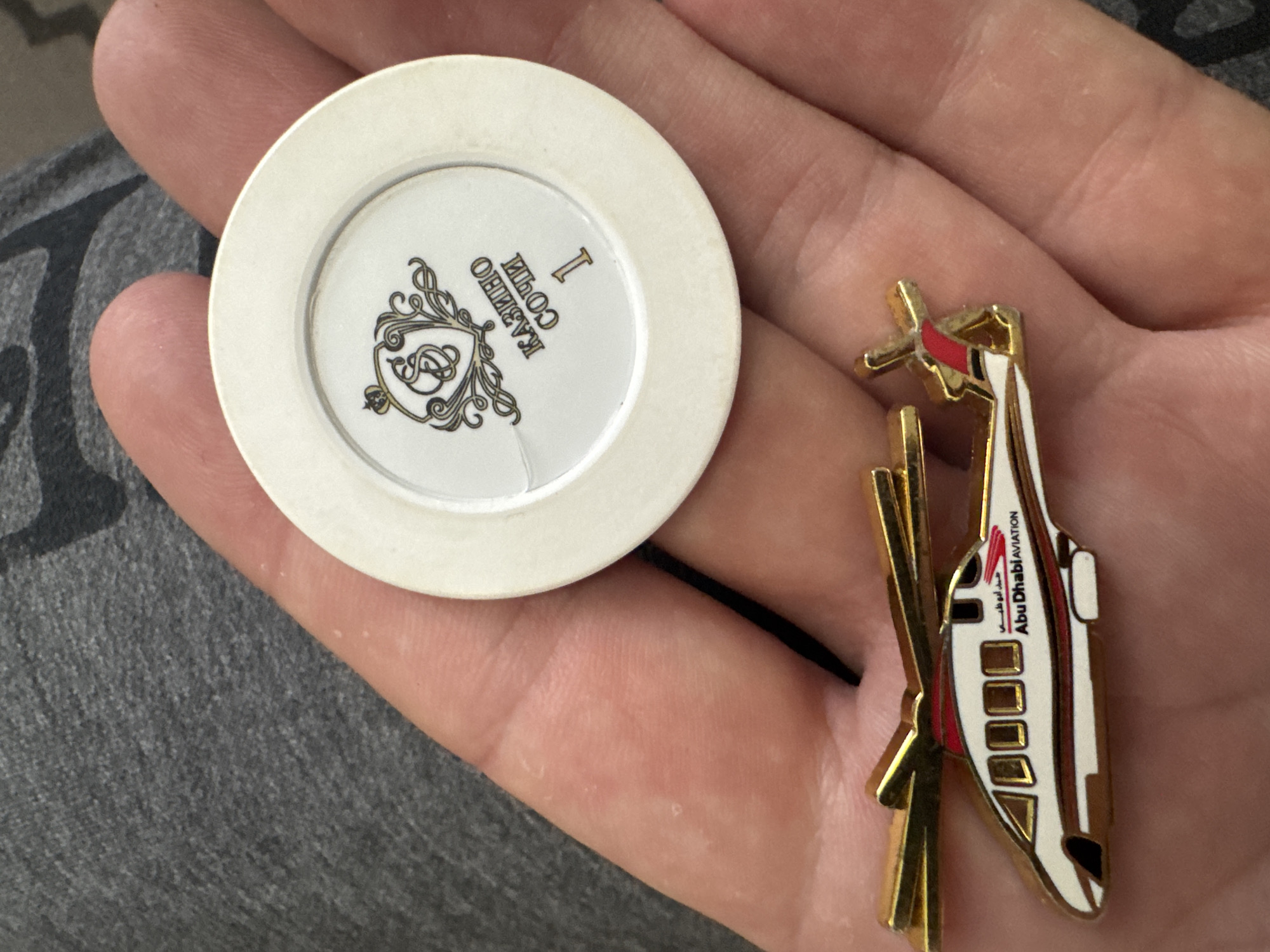 A Caucasian human hand holds a casino chip and a pin. The casino chip is white with a circular inset containing an ornate design, possibly a letter, and the words "КАЗИНО СОЧИ I" (Casino Sochi I) in Russian. The pin is a depiction of a white, gold, and red helicopter with "Abu Dhabi Aviation" printed on it. The hand is resting on a surface covered in grey fabric. The location is not explicitly mentioned as being in Chuprino, Russia.