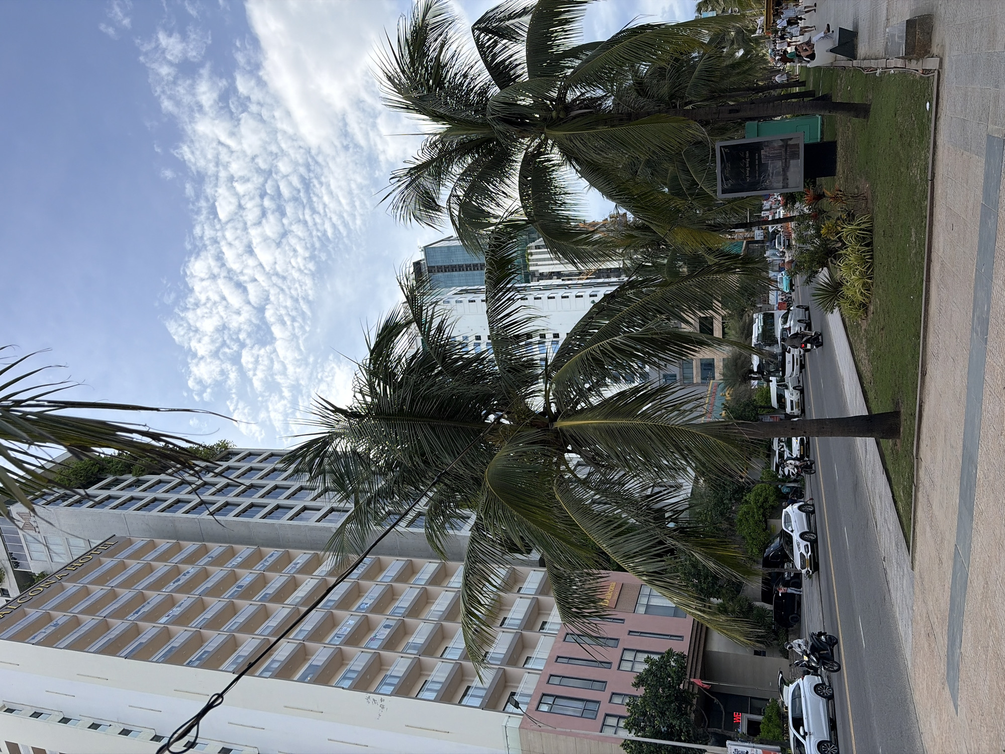 The image shows a street scene in Đà Nẵng, Vietnam, on a cloudy day. A multi-lane road runs parallel to the ocean, with a sidewalk on the right side. Several cars and motorcycles are on the road, and pedestrians are walking on the sidewalk. Palm trees line the sidewalk, and a few buildings are visible in the background, including a hotel. A large billboard is also present on the sidewalk.