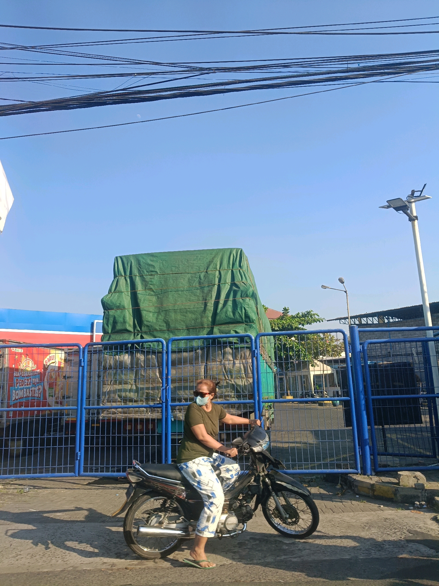 The image shows a woman wearing a green shirt, white and blue patterned pants, and a face mask, riding a black motorcycle. She is wearing sunglasses and green flip-flops. The motorcycle is facing towards the right of the frame. The woman is positioned in the lower right quadrant of the image. Behind her, a blue metal fence encloses an area with a large truck covered in a green tarp. The truck appears to be parked, and part of its cargo is visible, including colorful advertisements on the side. The background shows a clear blue sky with some power lines stretched across the top of the frame and a street lamp on the right. The scene appears to be outdoors, possibly near a port or industrial area in Surabaya, Indonesia, given the presence of a large truck and shipping containers, common in port cities. The lighting suggests it is daytime, with shadows cast on the ground.