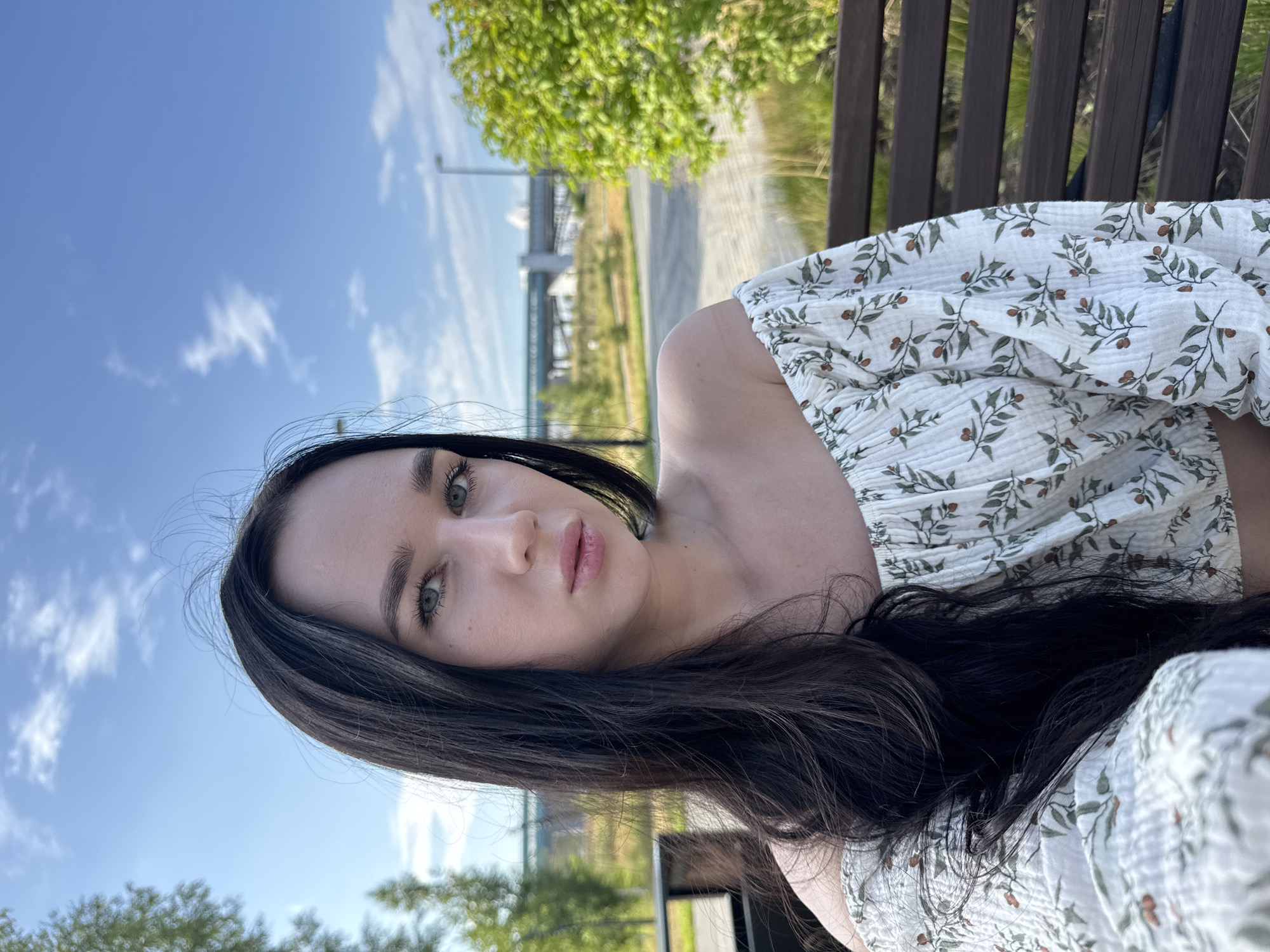 A young woman with long, dark hair and light-colored eyes is taking a selfie outdoors. She is wearing an off-the-shoulder white top with a floral pattern. The background shows a clear blue sky with some clouds, trees, and a street. The woman is sitting on a park bench. The scene appears to be in a public park or recreational area within the city of Novosibirsk, Russia.