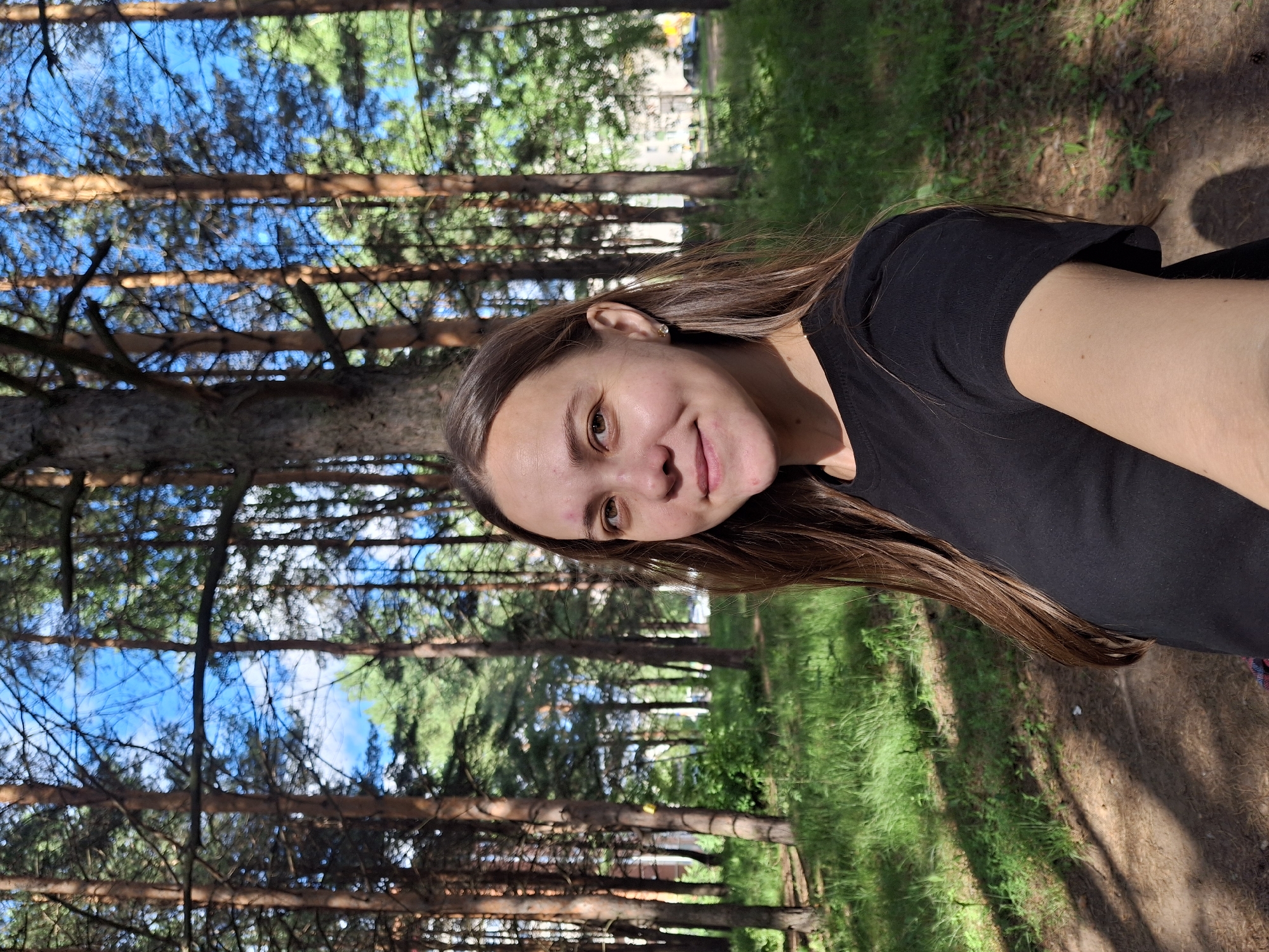 In a sun-dappled pine forest, a young woman with long brown hair and fair skin takes a selfie. She wears a black t-shirt and a small stud earring in her visible ear. Behind her, the tall, slender trunks of pine trees create a dense backdrop, with patches of bright blue sky visible through the canopy. Lush green grass and undergrowth cover the forest floor, with sunlight casting distinct shadows. In the distance, through a gap in the trees, a multi-story building and cars suggest the edge of an urban area, likely a residential district within the city of Chaikovsky, Russia. The overall scene conveys a sense of a warm, sunny day spent outdoors.