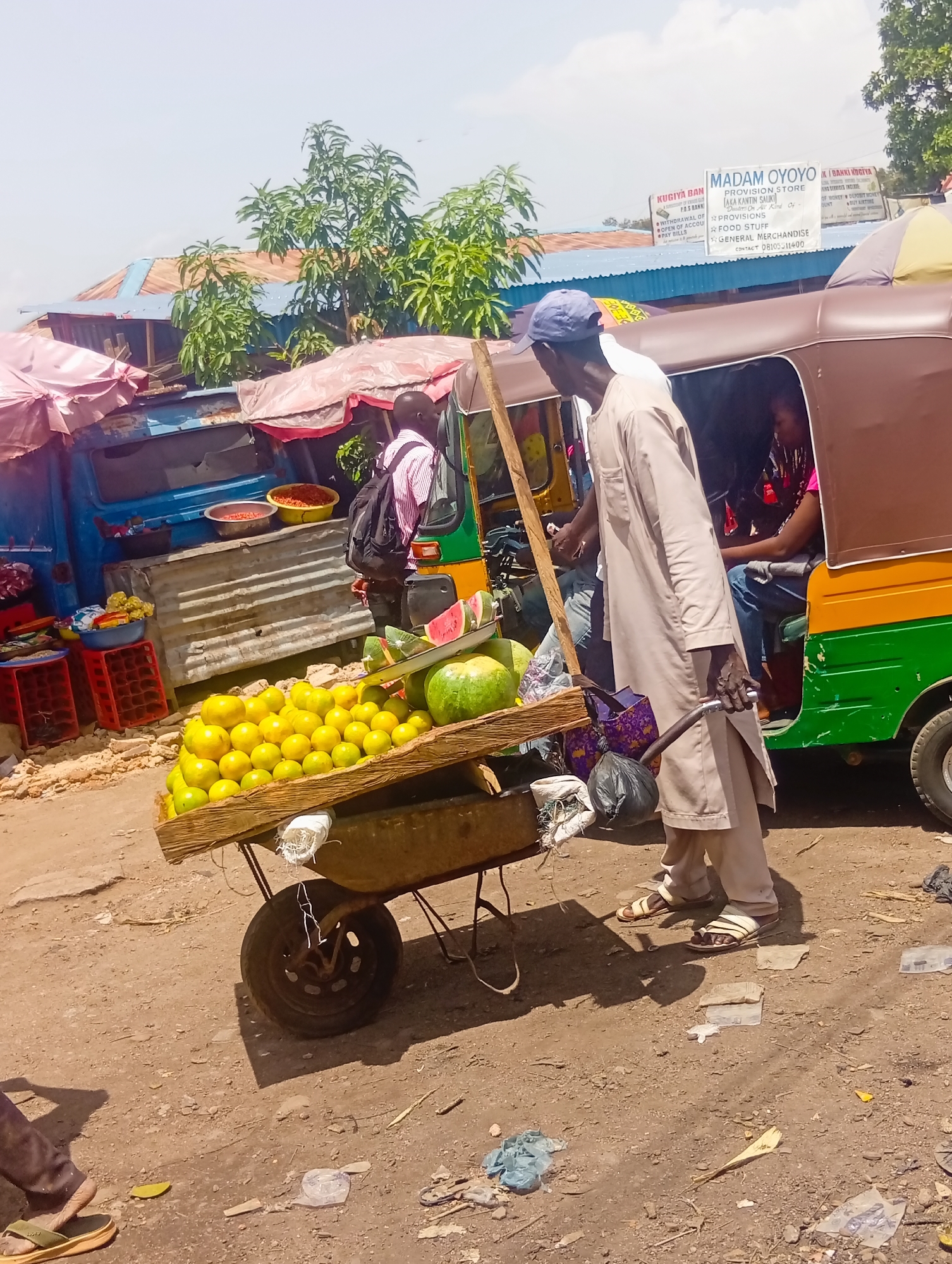 In Bukuru, Nigeria, a man wearing a light-colored kaftan and sandals stands beside a fruit cart loaded with a large quantity of yellow citrus fruits, possibly oranges or lemons, and several watermelons. The cart, a wooden platform on a single wheel, is positioned on a dirt road. Behind the cart, a three-wheeled auto-rickshaw, colored green and orange, is parked. A person wearing a pink top is visible inside the auto-rickshaw. The backdrop consists of stalls with corrugated metal roofing, some covered with pink tarps, and trees. Signs with text in English are visible on some buildings in the background, including one that reads "MADAM OYOYO PROVISION STORE". The scene appears to be an outdoor market.