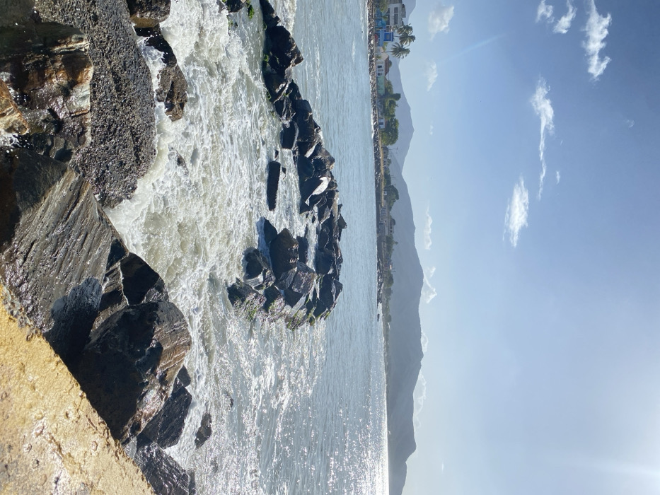 Waves crash against dark, jagged rocks along a coastline. In the distance, a line of buildings sits below a mountain range. The sky is bright blue with scattered wisps of clouds. The water reflects the sunlight, creating a shimmering effect.