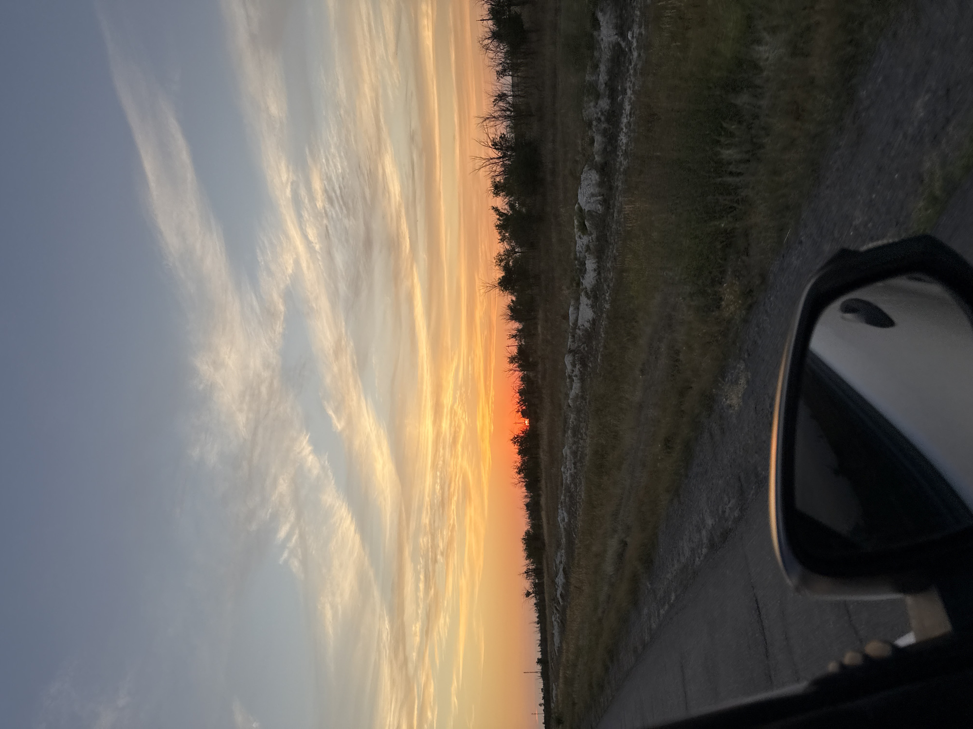 The image captures a view of a sunset over a rural landscape, as seen from inside a vehicle. The sky is painted with warm hues of orange and yellow near the horizon, transitioning into lighter shades of pink and blue towards the top, with wispy clouds streaking across. A line of trees and vegetation borders the field, silhouetted against the vibrant sky. In the foreground, the side-view mirror of the vehicle reflects the sky and the surrounding environment. A portion of the road and the grassy roadside are visible to the left of the mirror. The scene suggests a journey through a sparsely populated area. The location is identified as Pugachyov Rural District, Kazakhstan, which aligns with the rural setting.
