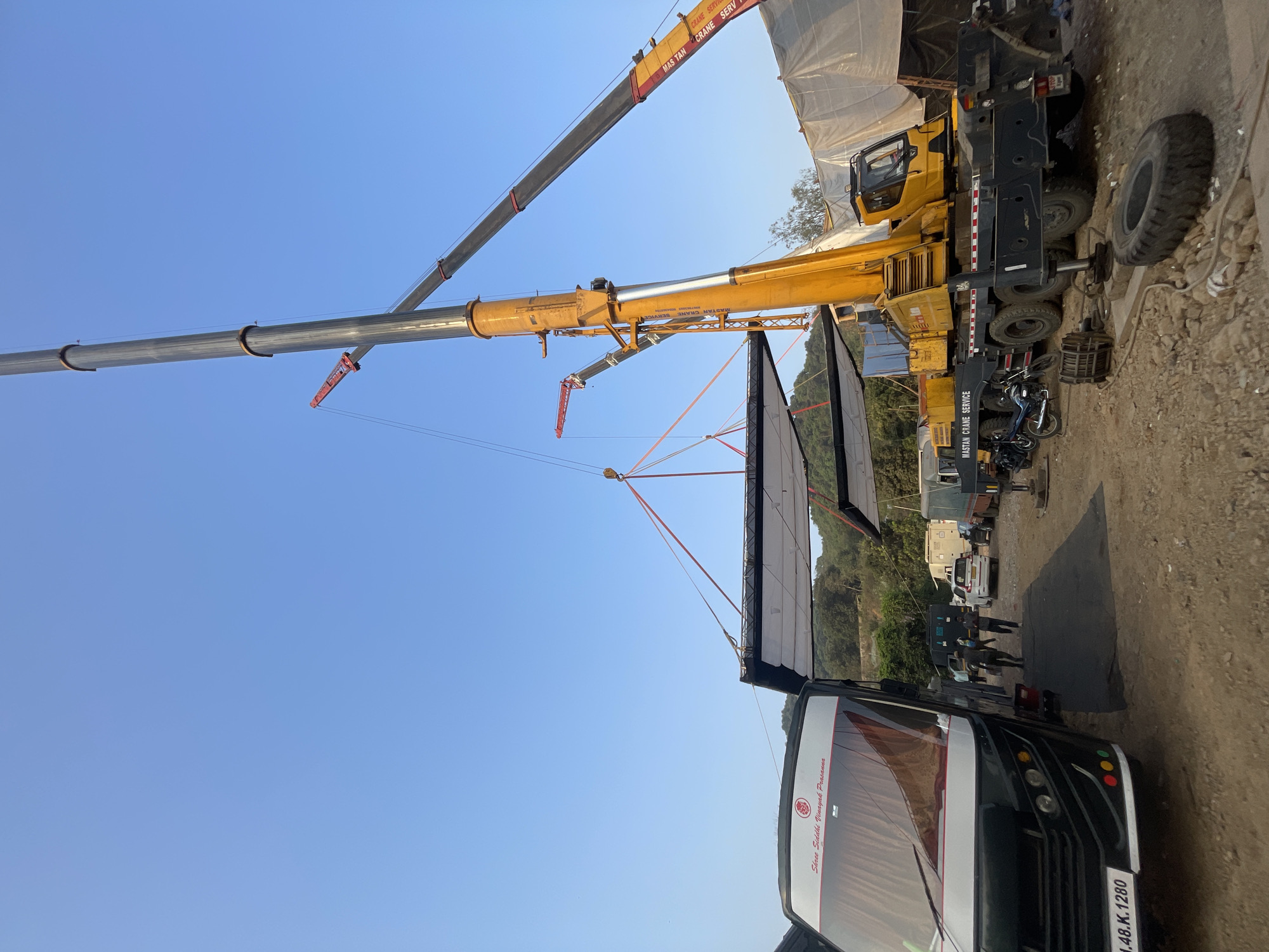 The scene depicts a bustling film set in what appears to be Mira-Bhayander, India, indicated by the license plate "1.48.K.1280" on a black and white bus labeled "Shree Siddhi Vinayak Prasanna," which is common in the area. A massive crane, branded "MASTAN CRANE SERVICE," dominates the view, extending its long, silver boom towards a large, rectangular screen being hoisted into place. This setup suggests an outdoor film shoot involving large-scale equipment. People are visible in the background, likely crew members involved in the production. The terrain is uneven and somewhat dusty, typical of film set environments. Other vehicles are parked nearby, indicating a sizable crew presence.