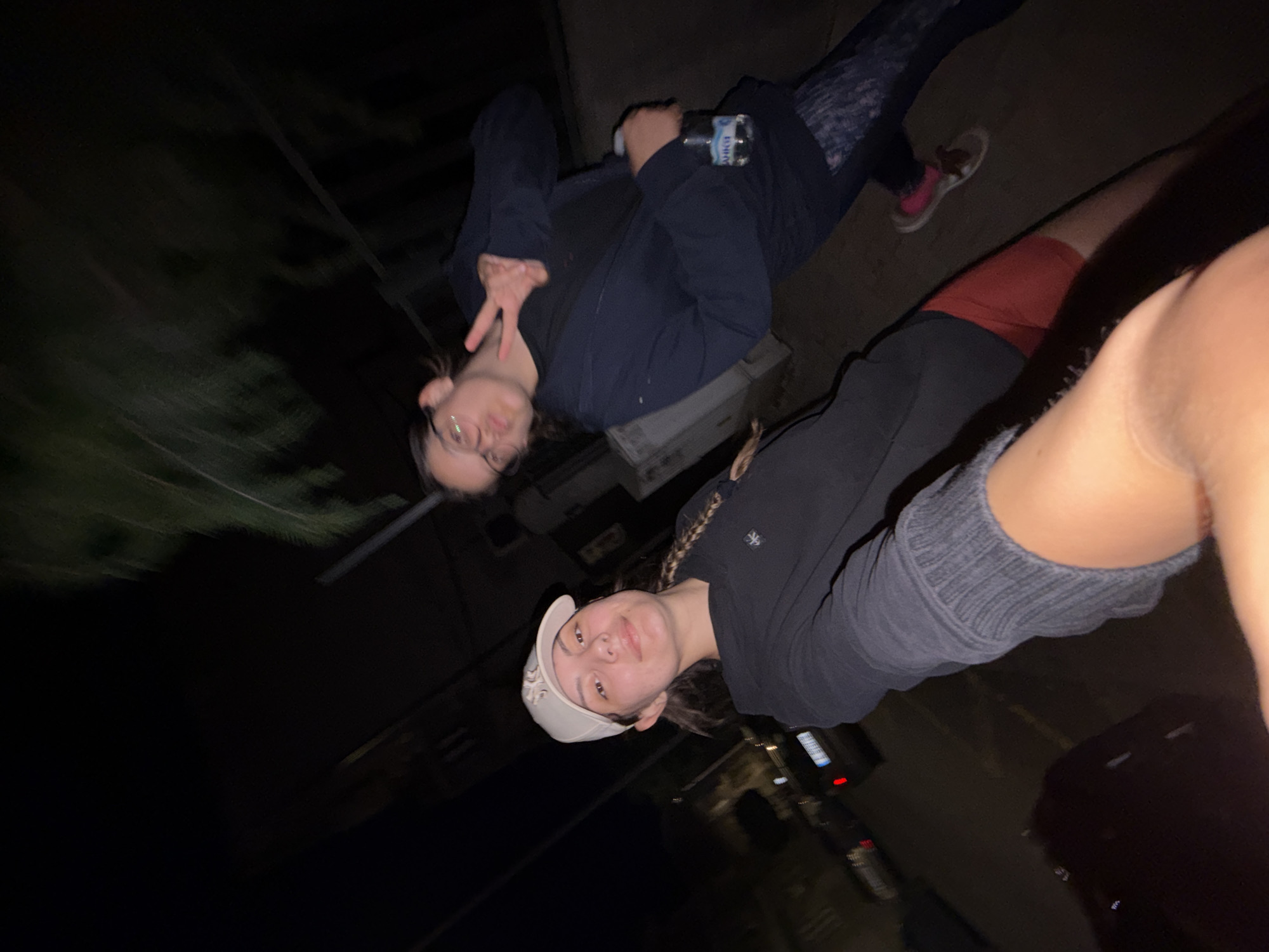 The image depicts two individuals at night in a city. One person in the foreground wears a white baseball cap and a black hoodie. Another person in the background wears glasses and a dark jacket, holding a water bottle and making a peace sign. The environment appears to be an outdoor urban setting, possibly a street or sidewalk, with some greenery visible. The overall lighting is dim, indicating it is nighttime. The city of Burgas, Bulgaria is not explicitly identifiable from the visual information within the image.