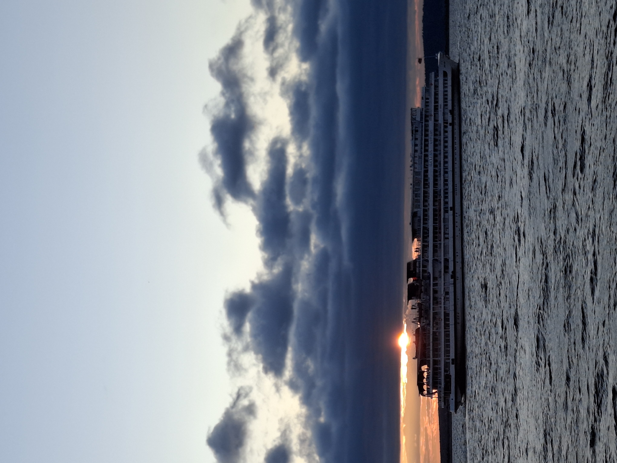 A wide-angle shot captures a large, multi-decked white cruise ship sailing on dark, choppy water under a dramatic sky. The sun is setting, partially obscured by dark, heavy clouds, casting a warm orange glow along the horizon. The ship, positioned in the center of the frame, displays numerous windows and balconies on its multiple levels. Silhouettes of people can be seen on the upper decks. The water is rippled, reflecting the dim light of the setting sun and the dark sky. The overall scene suggests a tranquil yet imposing maritime setting at dusk.