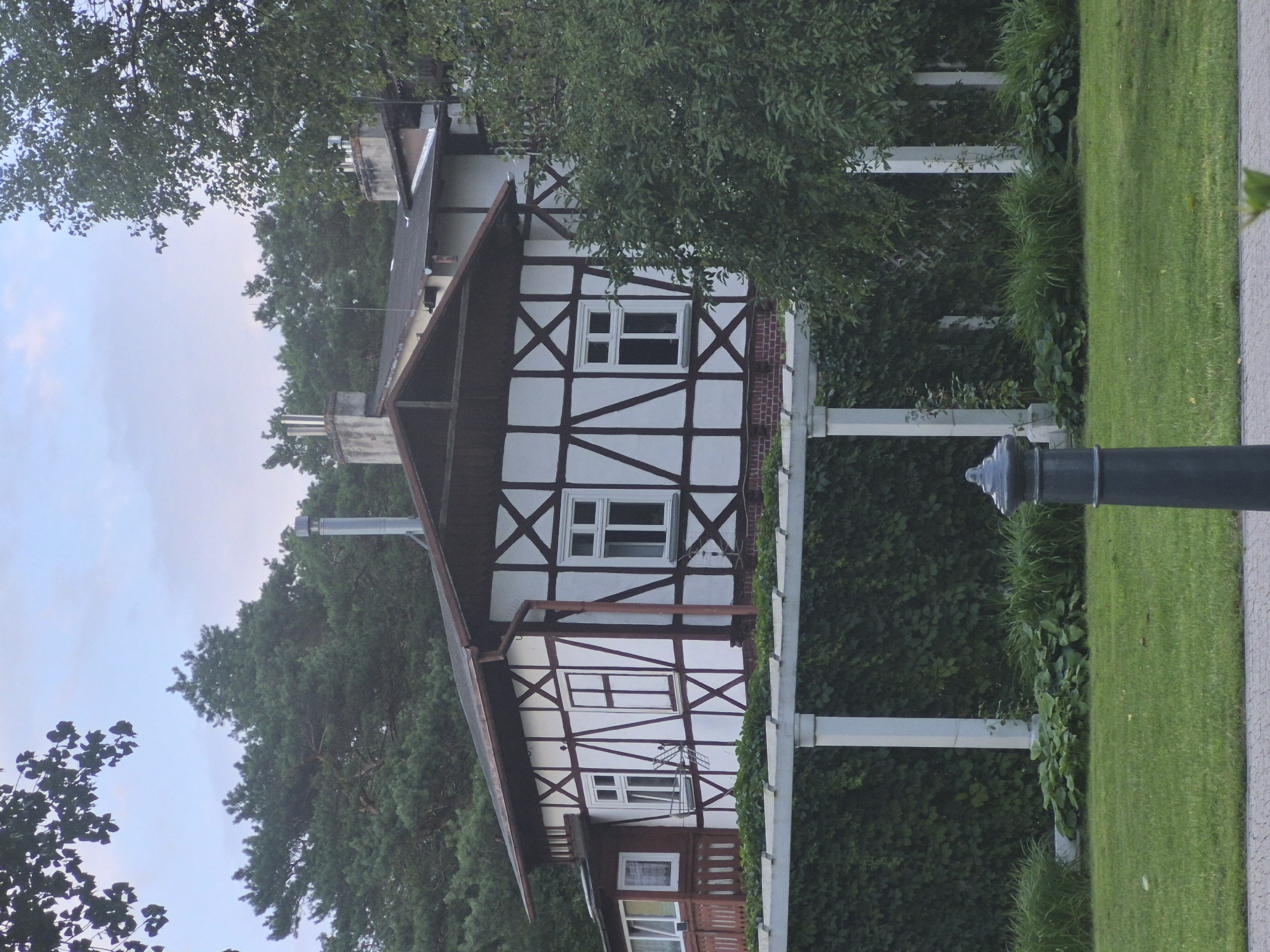 The image captures a two-story building with a distinctive half-timbered facade, featuring white infill panels and dark timber framing. The roof is dark brown with visible gutters. Several windows with white frames are visible on the facade. A portion of a balcony with a red railing is also present on the left side of the building. The building is situated behind a white pergola structure supported by pillars, which is covered in dense green ivy. In front of the pergola, there is a neatly maintained green lawn. A dark grey lamppost with a decorative top stands in the foreground on the right side of the image, and another lamppost is visible on the far left edge of the frame. The background consists of a dense cluster of green trees. The sky is overcast and light blue. The scene appears to be set in a park or garden area, likely a residential or public building. This particular architectural style is common in Gdańsk, Poland, especially in areas with historical German influence, suggesting it could be a villa or a historical building within the city.