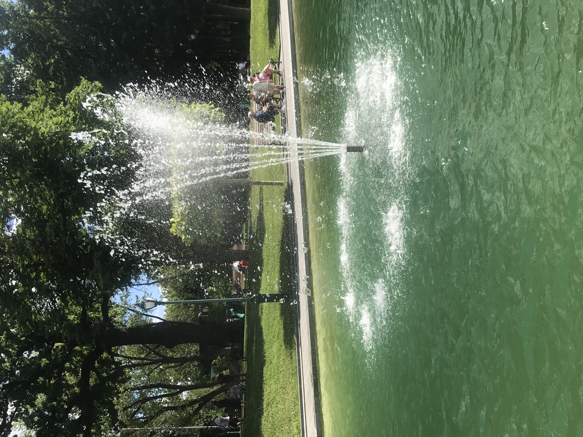 This image depicts a park scene in Kharkiv, Ukraine, featuring a fountain in a green pond. The fountain's spray catches the sunlight, creating a sparkling effect. In the background, people are seated on benches on a grassy incline, shaded by large trees. A paved walkway runs along the edge of the pond. The water in the pond is a vibrant green, and the surrounding environment suggests a sunny day.