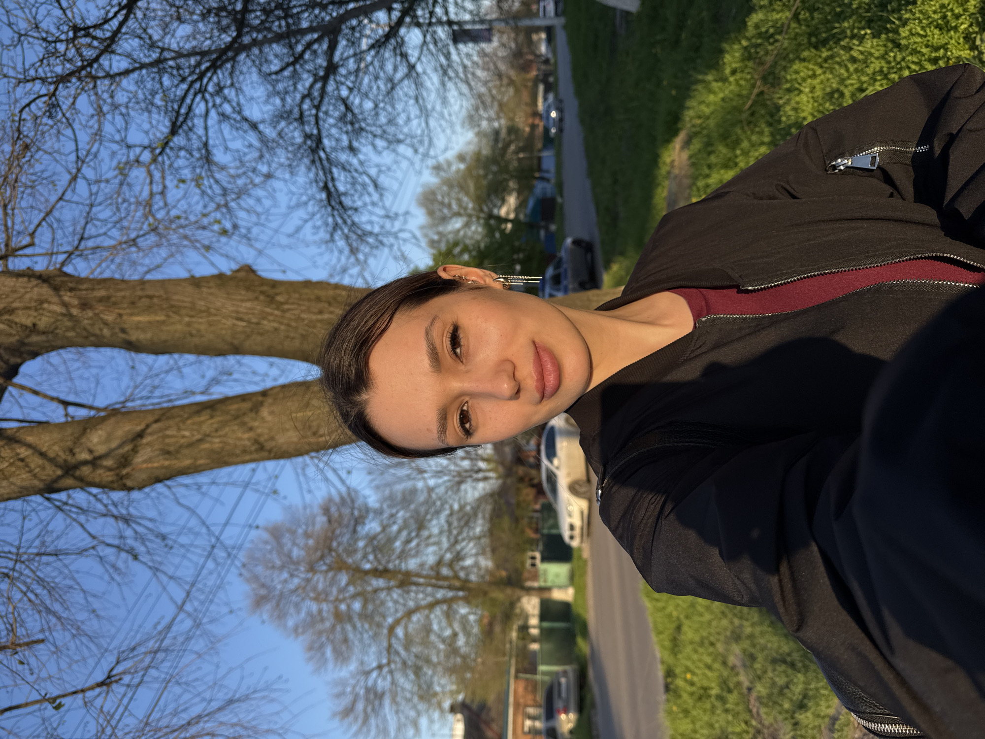 A person is posing for a selfie outdoors. The individual has brown hair pulled back, fair skin, and is wearing a black jacket over a dark red shirt. They are facing the camera with a slight smile. The background shows a residential street with houses, parked cars, trees, and a sidewalk. The sky is blue, and sunlight is illuminating the scene.