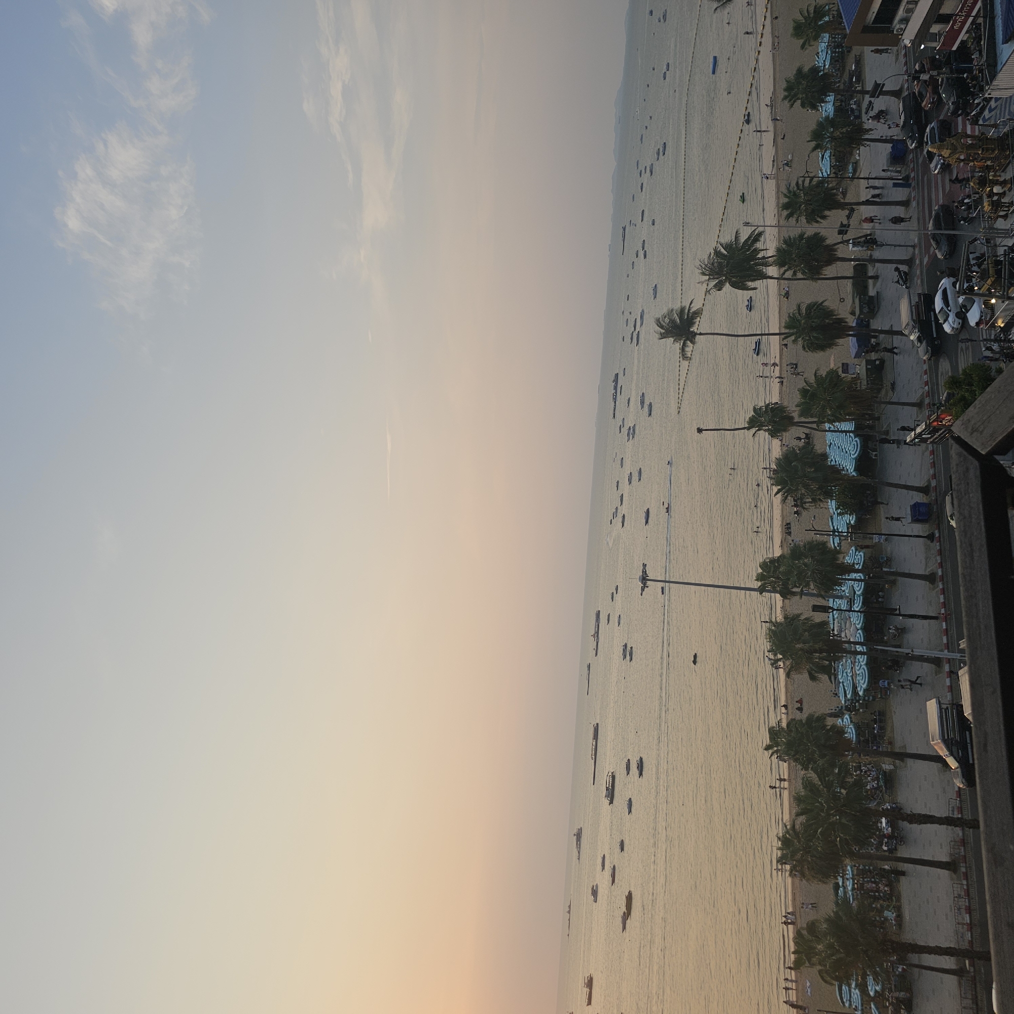 The image depicts a high-angle view of Pattaya Beach in Pattaya City, Thailand, likely taken from a building. The scene is divided between the sky on the left and the beach/water on the right. The sky transitions from a light blue at the top to a warmer, orange-tinted hue near the horizon.
The beach stretches along the coastline. The water is relatively calm, with numerous boats visible scattered across the surface. Along the shoreline, there are palm trees lining the beach. Light-colored, circular objects (likely beach umbrellas) are arranged in rows on the sand. A road runs parallel to the beach, and there are buildings situated alongside the road. The quality of the image suggests it was taken during the evening. The details of the street and people are clear enough to notice there are vehicles on the street and people are on the beach.