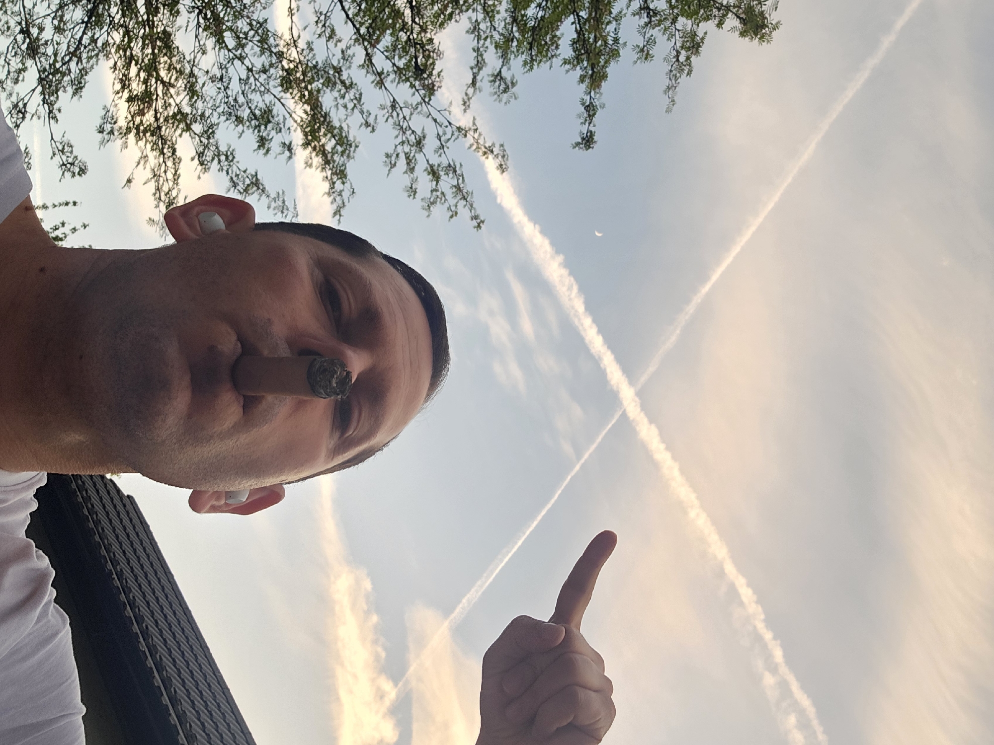 A man holds a lit cigar in his mouth, points with his right index finger towards the sky, and has a white in-ear device in his left ear. The sky is visible and crossed by two vapor trails, with a faint crescent moon. A leafy tree is visible at the top of the frame. A dark object, possibly part of a building's structure, is partially visible on the bottom left. The location in London is not discernible from the image.