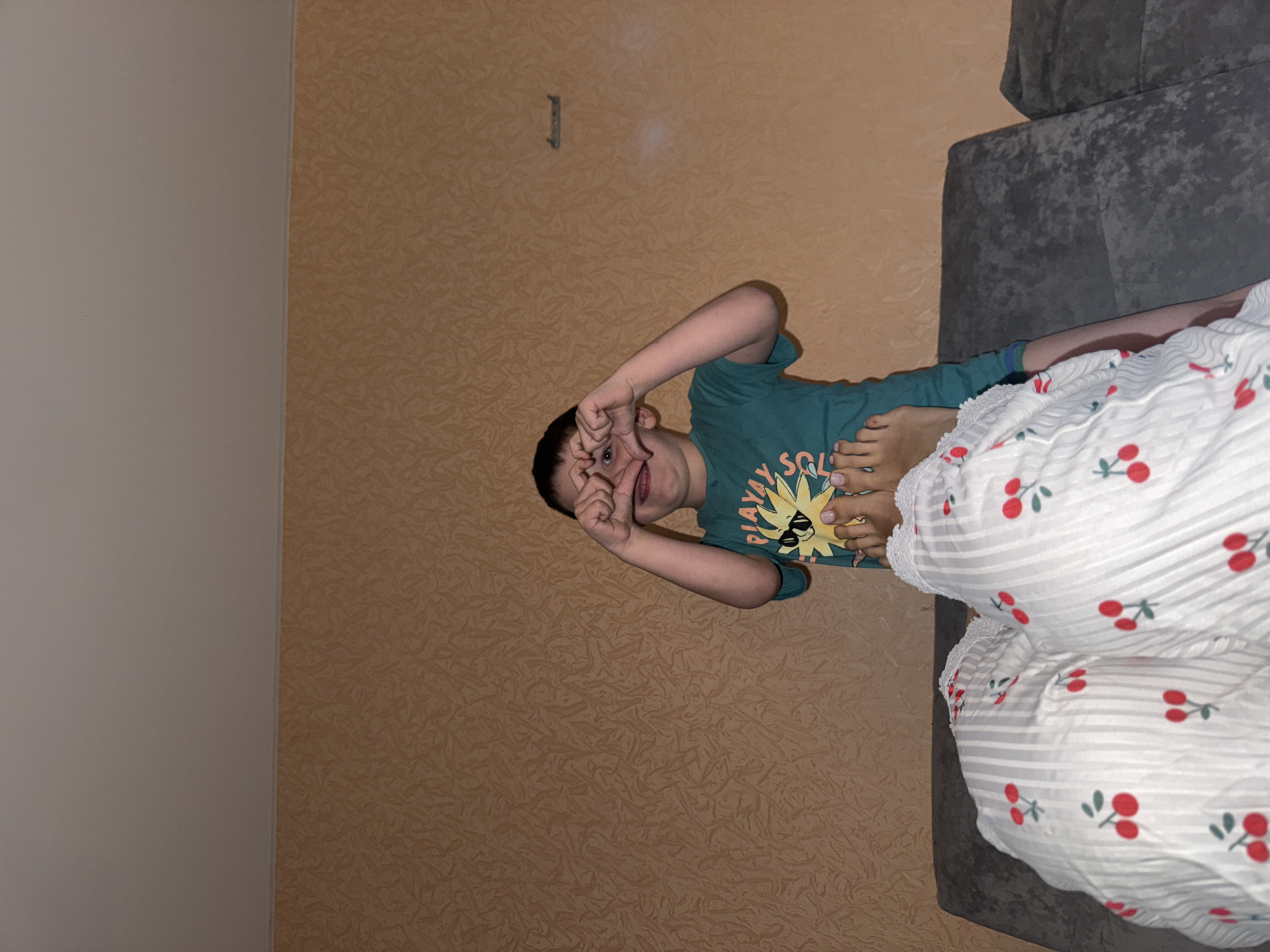 This indoor image, set in Chaikovsky, Russia, captures a playful moment with a young boy and an adult. The boy, positioned towards the center-right of the frame, is lying down and wearing a teal-green t-shirt. His shirt features a yellow sun graphic with the visible text "AY SOL" and other indistinct words. He has dark hair and is looking directly at the camera, forming a heart shape with his hands over his eyes, with an open mouth suggesting a smile or laugh.
To the right, only the feet and lower legs of an adult are visible, covered by a white blanket featuring red cherry motifs with green stems and grey stripes. The adult's toenails are painted a light color. A grey, upholstered sofa or couch is partially visible behind the blanket. The wall in the background is covered with textured, light brown or beige wallpaper. A small, dark, rectangular object or mark is visible on the upper portion of the wall. The scene conveys a cheerful and casual atmosphere under artificial indoor lighting.
