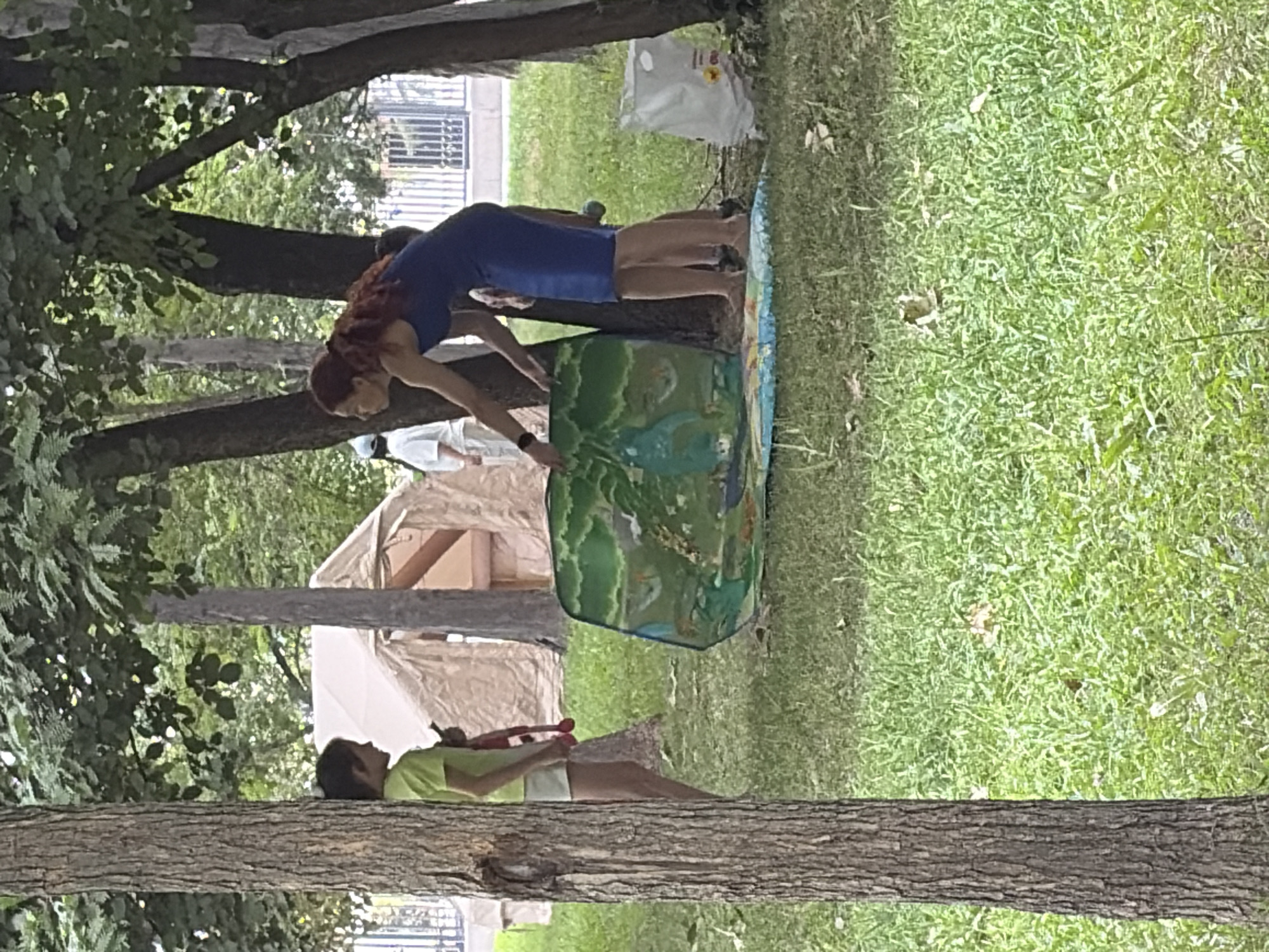 In a grassy park area with trees, a woman in a blue dress is placing a large, colorful playmat depicting a savanna scene on the ground. A child in a lime green shirt and white shorts stands nearby, holding a red toy. In the background, there are tents and a fence.