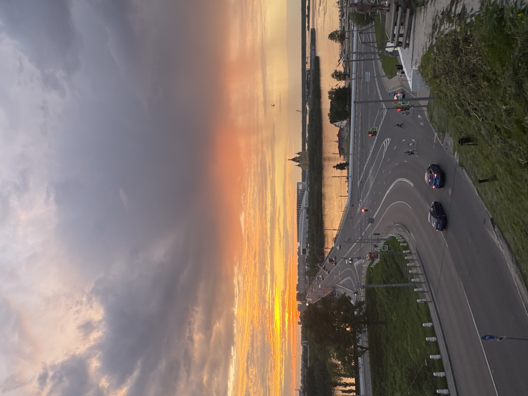 The image captures a sunset view of Nizhny Novgorod, Russia. The sky is divided between dramatic gray clouds on the left and vibrant orange and yellow hues on the right, reflecting the setting sun. The foreground features a road with several cars moving along it. There are pedestrian crossing signs, traffic lights, and streetlights alongside the road. The road curves, bordered by grass. In the distance is the Volga River and buildings. The Alexander Nevsky Cathedral can be seen on the horizon.